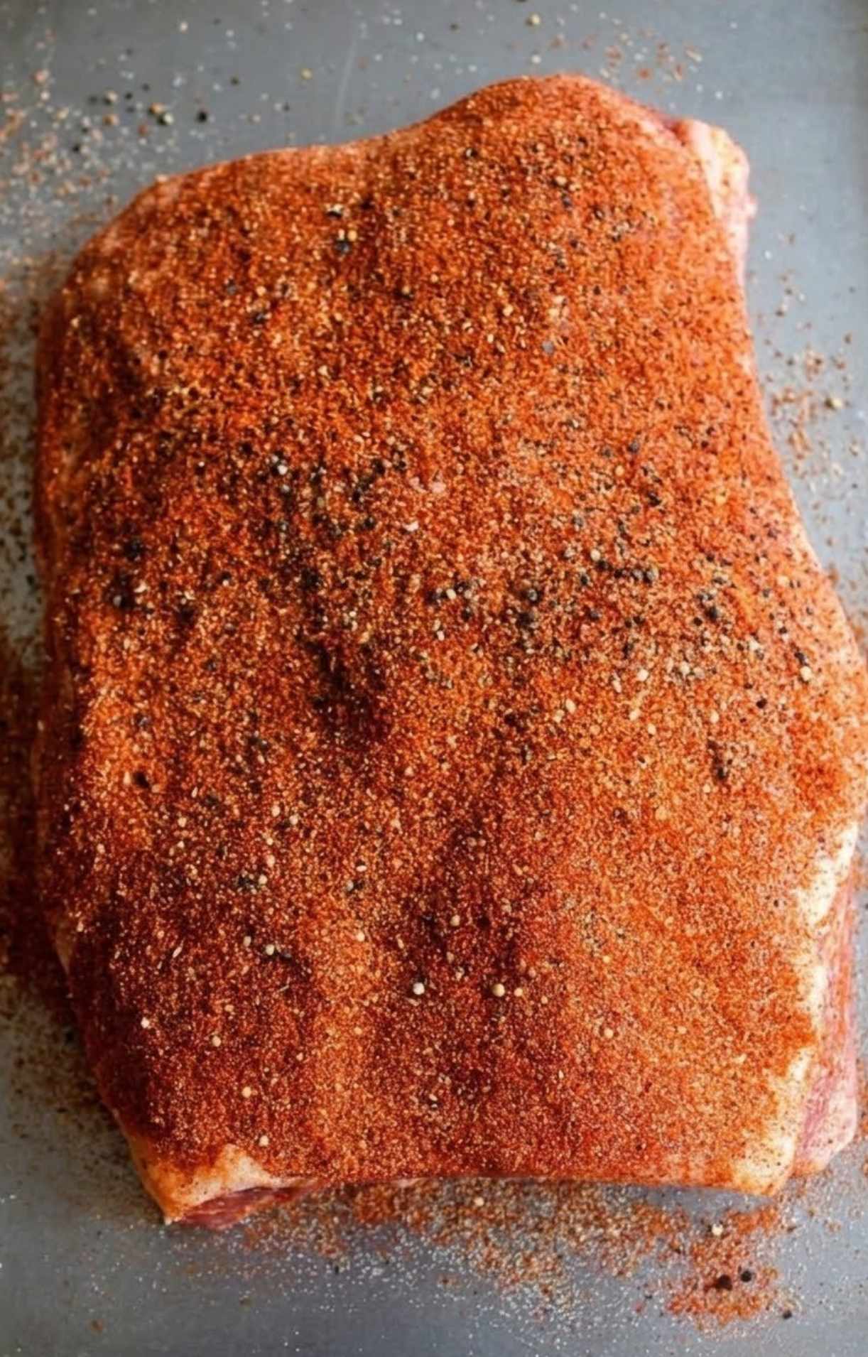 A large cut of raw beef brisket seasoned generously with a thick, even layer of smoked beef brisket rub on a metal baking sheet.