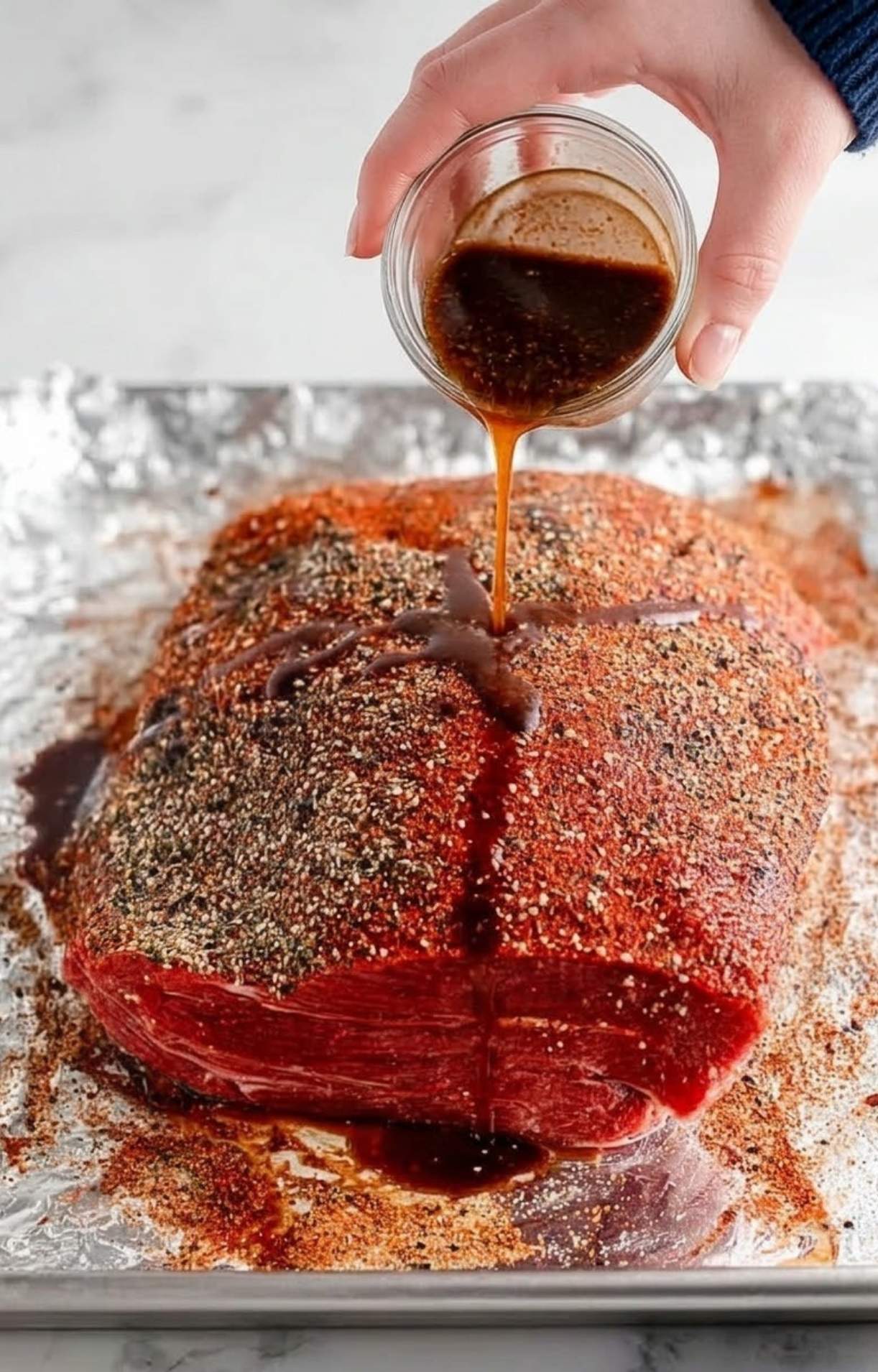 A hand pouring a dark liquid, likely liquid smoke or a marinade, over the seasoned raw beef brisket on a foil-lined baking sheet.