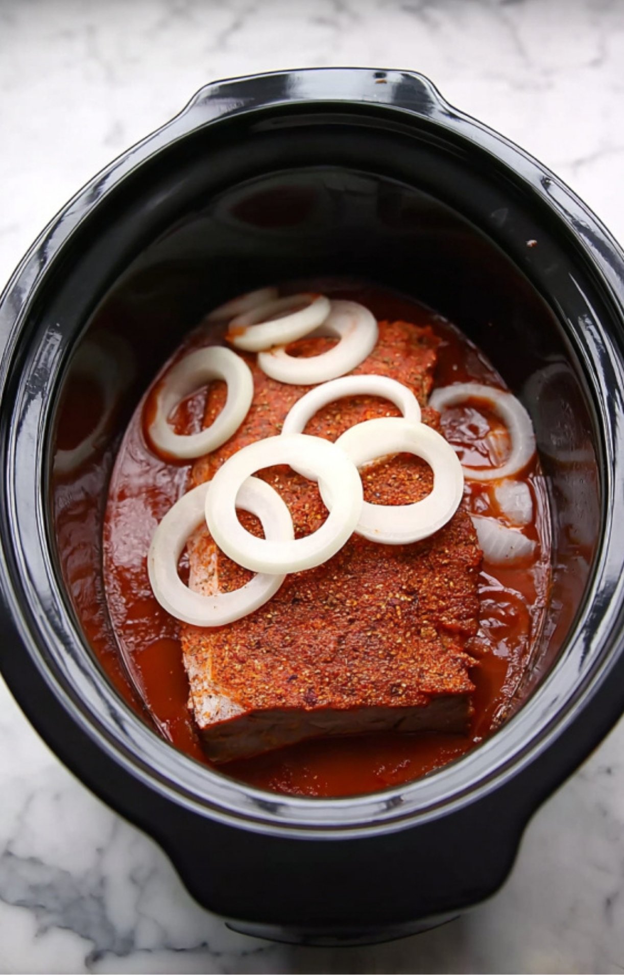 Seasoned beef brisket placed in a slow cooker topped with fresh onion rings and a rich barbecue base.