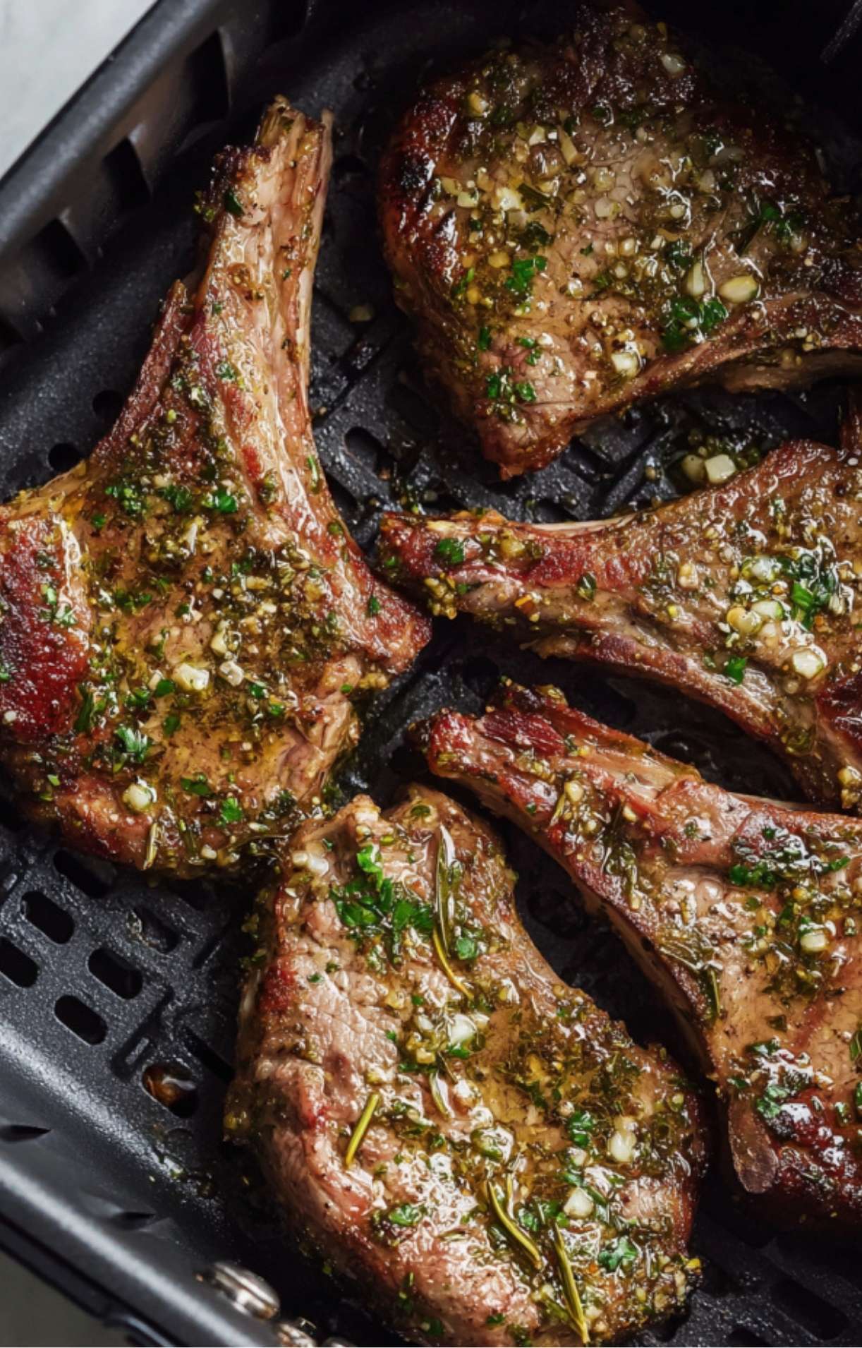 Top-down view of five lamb chops seasoned with fresh parsley and garlic cooking inside a black air fryer basket.
