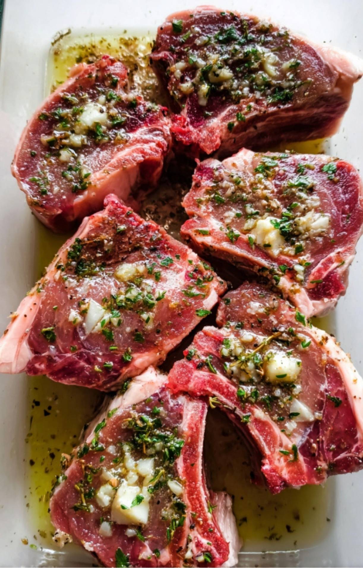 Raw lamb loin chops resting in a glass dish, generously coated with a garlic, herb, and olive oil marinade.