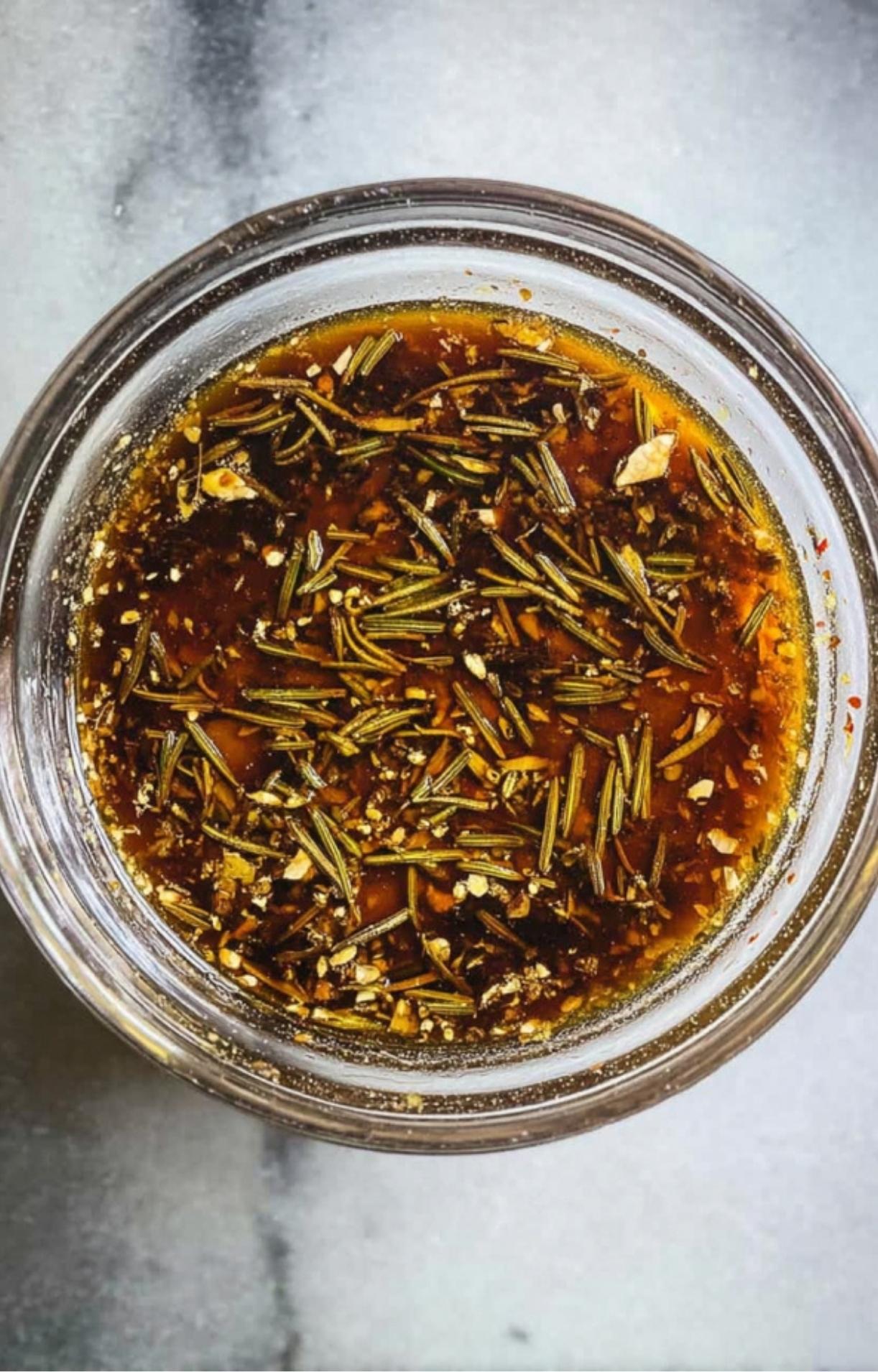 A glass bowl containing an oil-based marinade with dried rosemary and spices for air fryer lamb shoulder chops.