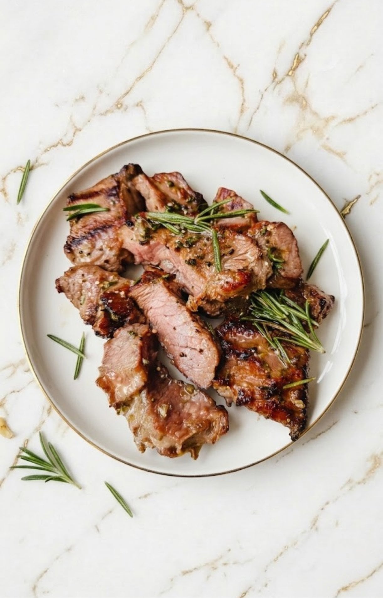 Sliced pieces of medium-cooked lamb steak garnished with fresh rosemary on a white plate with a gold rim.