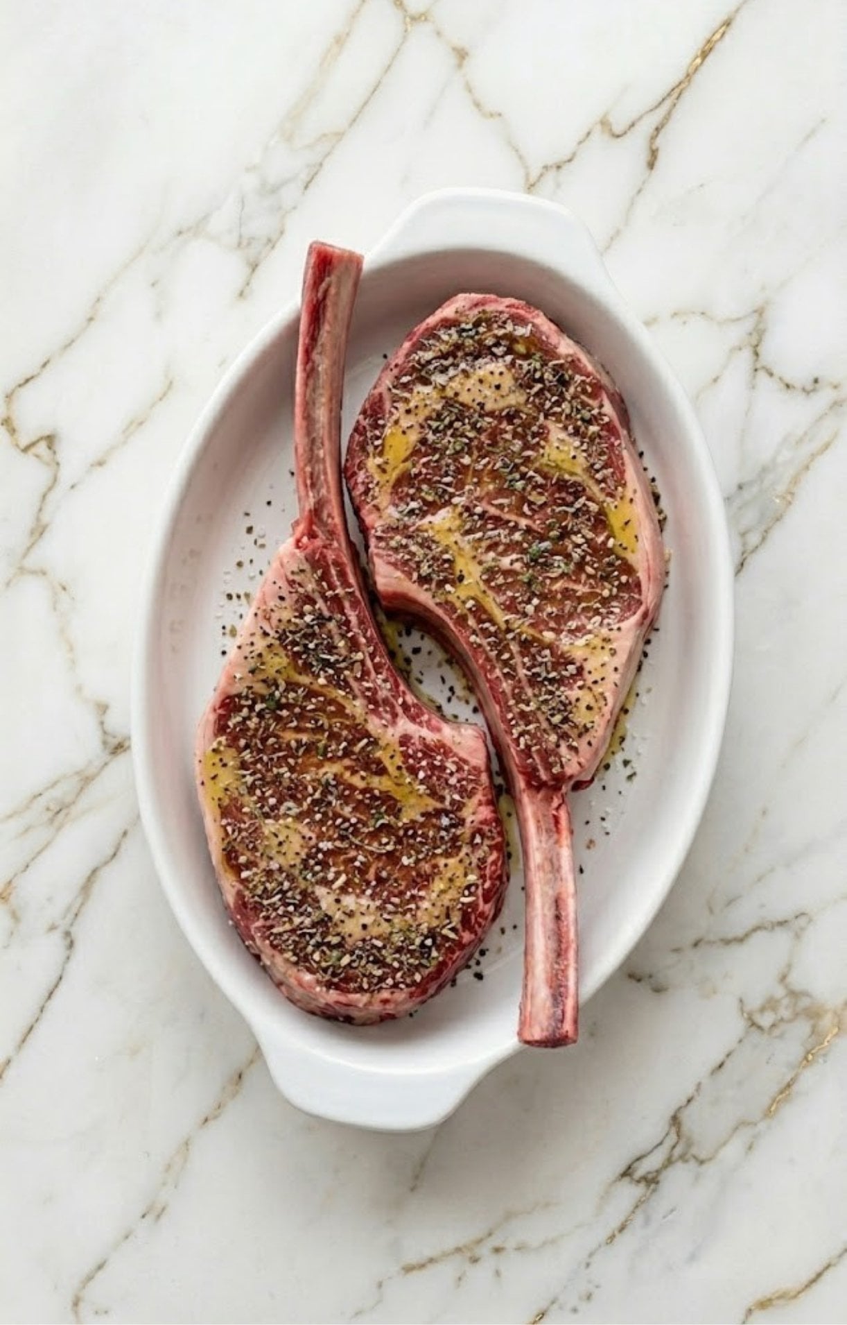 Raw tomahawk steaks in a white dish coated generously with a dark peppercorn and herb dry rub mixture over a light drizzle of olive oil.