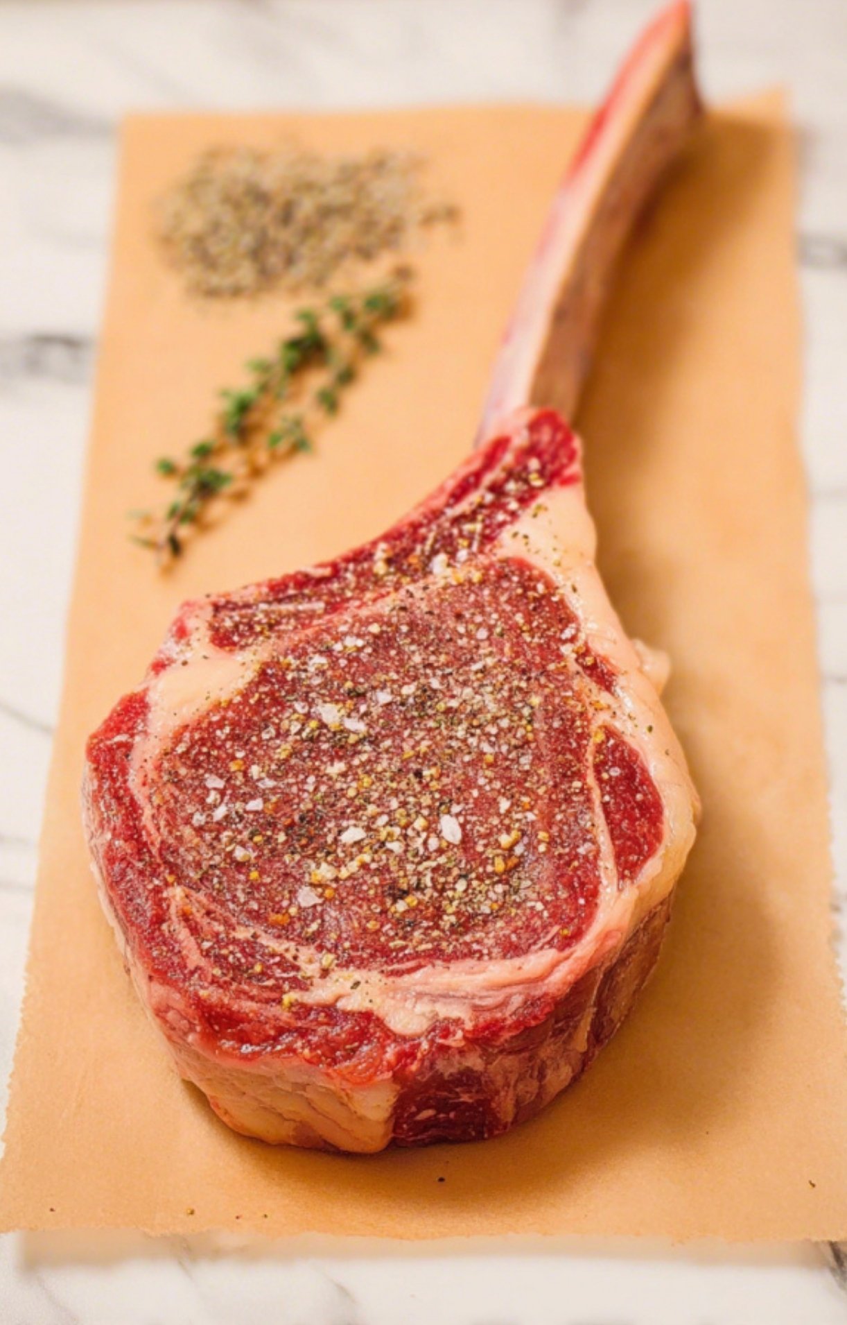 A thick, raw bone-in tomahawk steak seasoned generously with salt, pepper, and herbs on parchment paper.