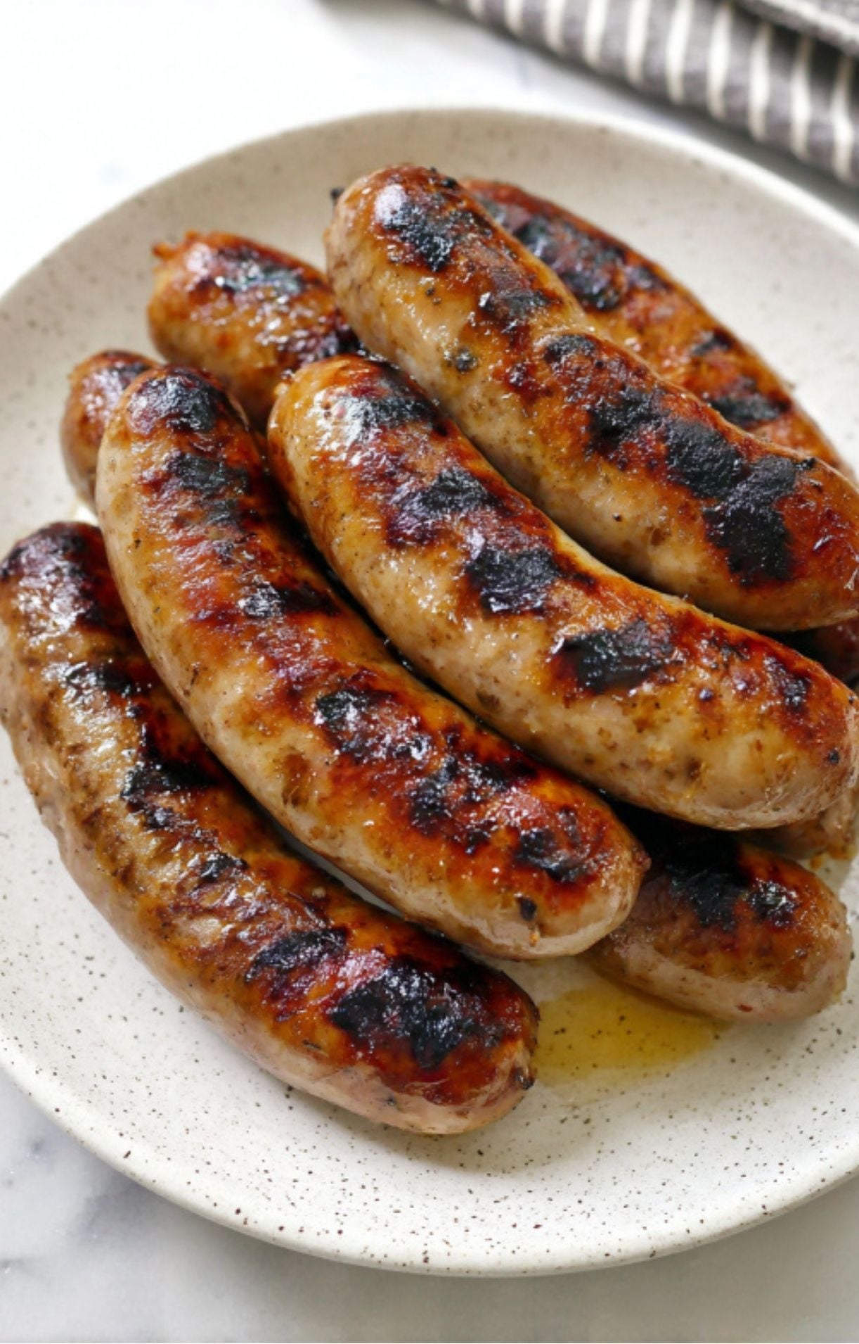 A pile of juicy, perfectly seared and charred beer-braised bratwurst sausages on a speckled white plate.