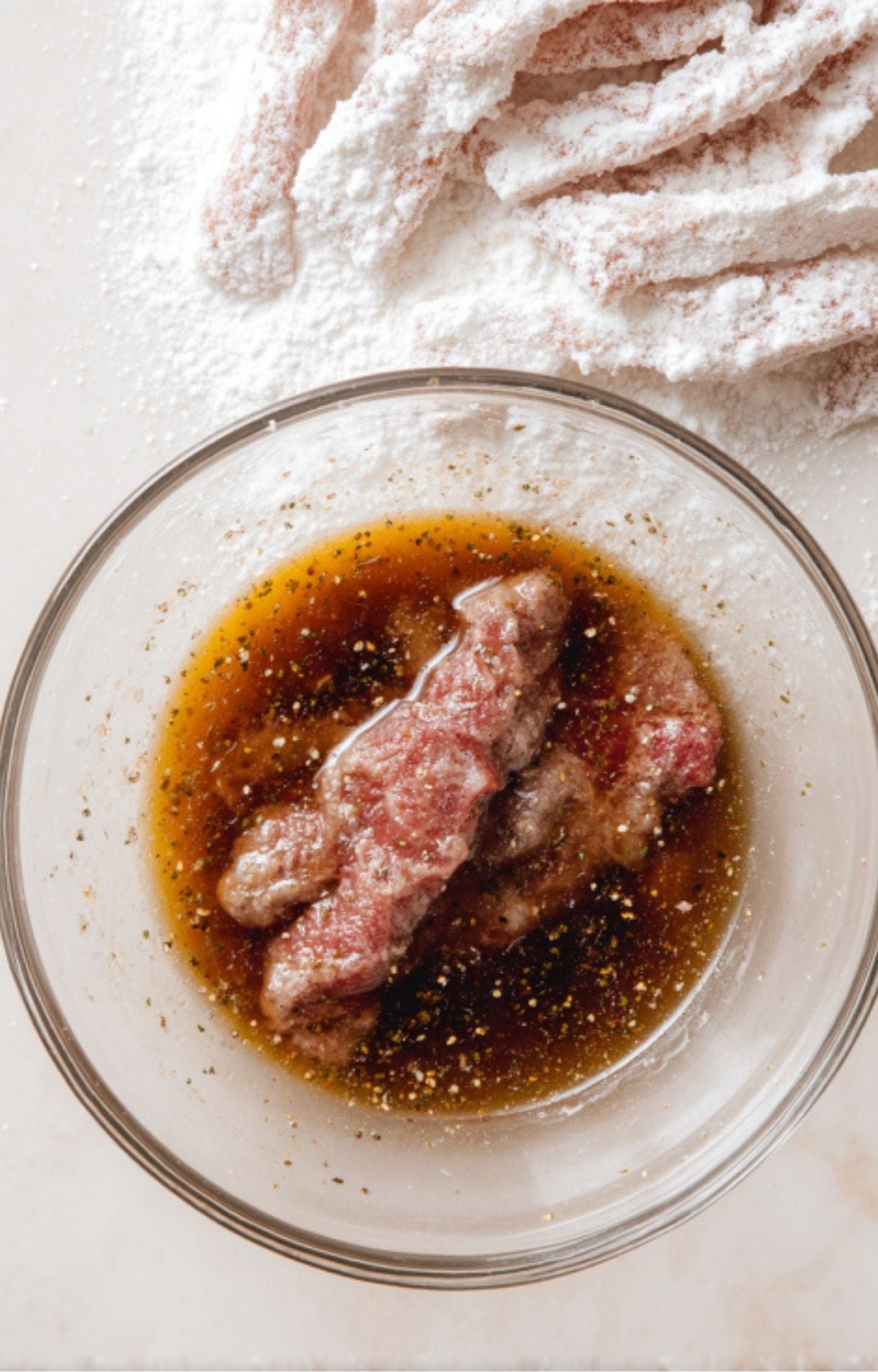 Slices of beef being dipped into a liquid marinade next to a pile of white cornstarch coating.