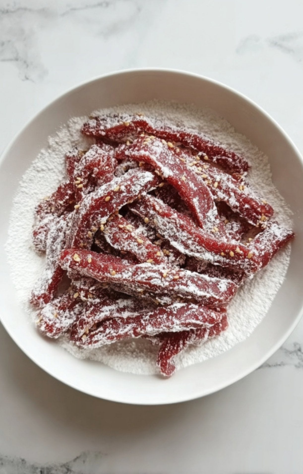 Thinly sliced raw beef strips coated in white cornstarch and sesame seeds in a bowl for a crispy Beijing Beef stir fry.