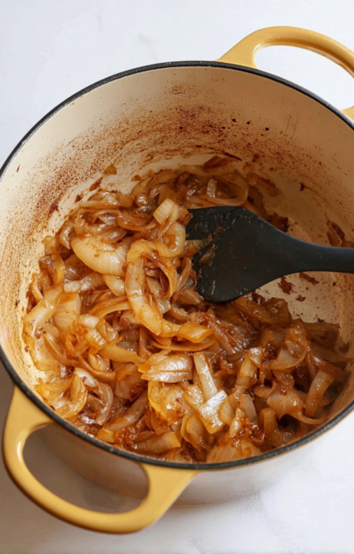Caramelized sliced onions being stirred with a black spatula in a yellow Dutch oven to create a flavorful base for Beer Boiled Brats.