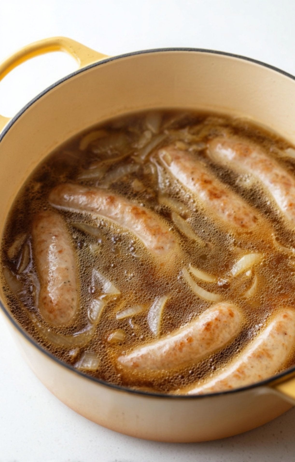 Raw bratwurst links submerged in a bubbling beer and onion broth inside a yellow pot, simmering to infuse the meat with flavor.