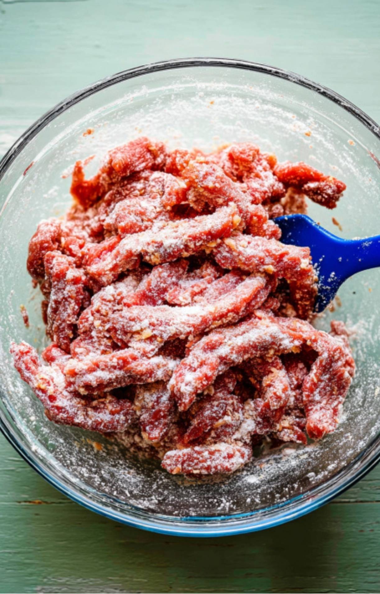 Raw beef strips coated in a white flour and cornstarch mixture being tossed with a blue spatula.