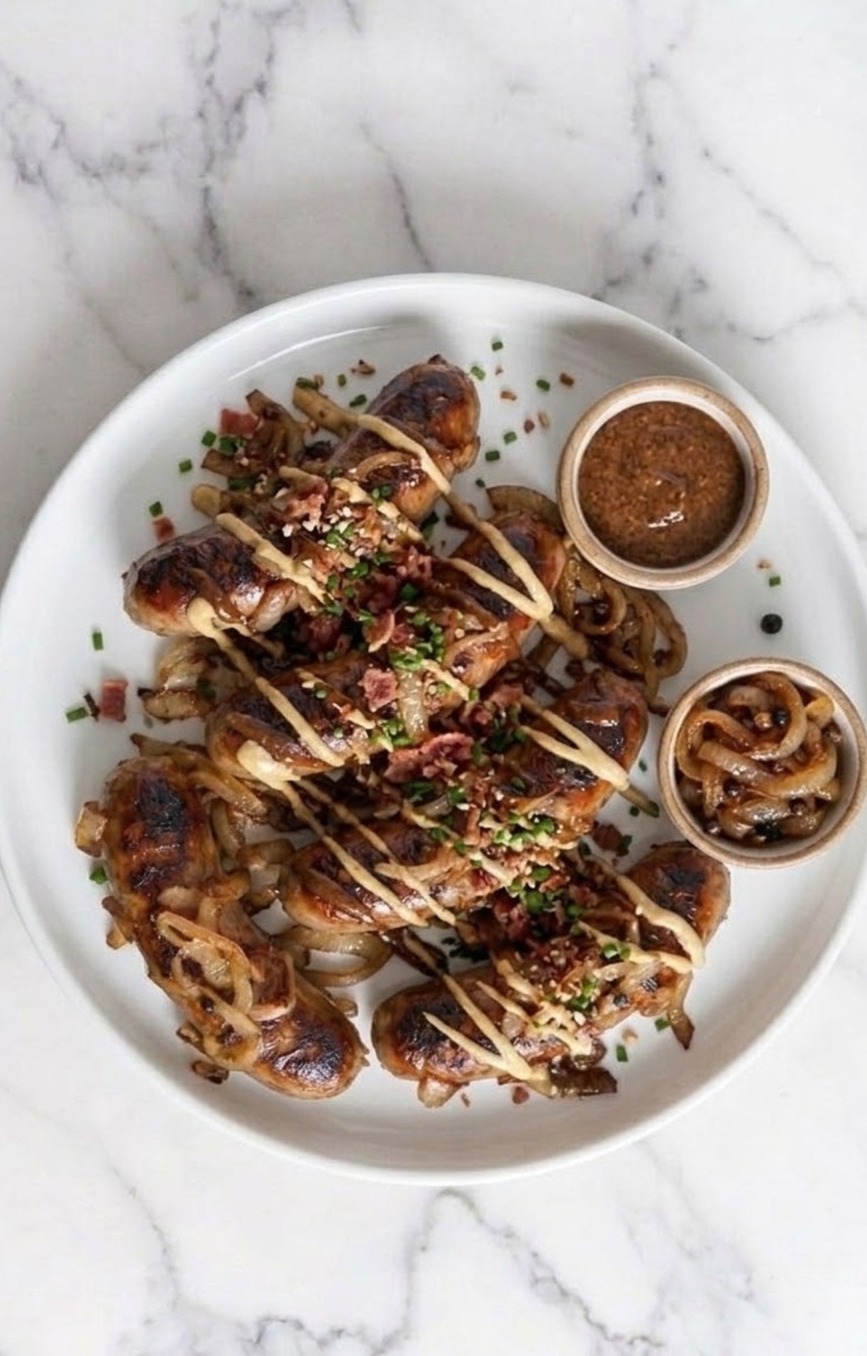 A white plate featuring grilled beer brats topped with onions, bacon bits, and chives, served with a side of grainy mustard.