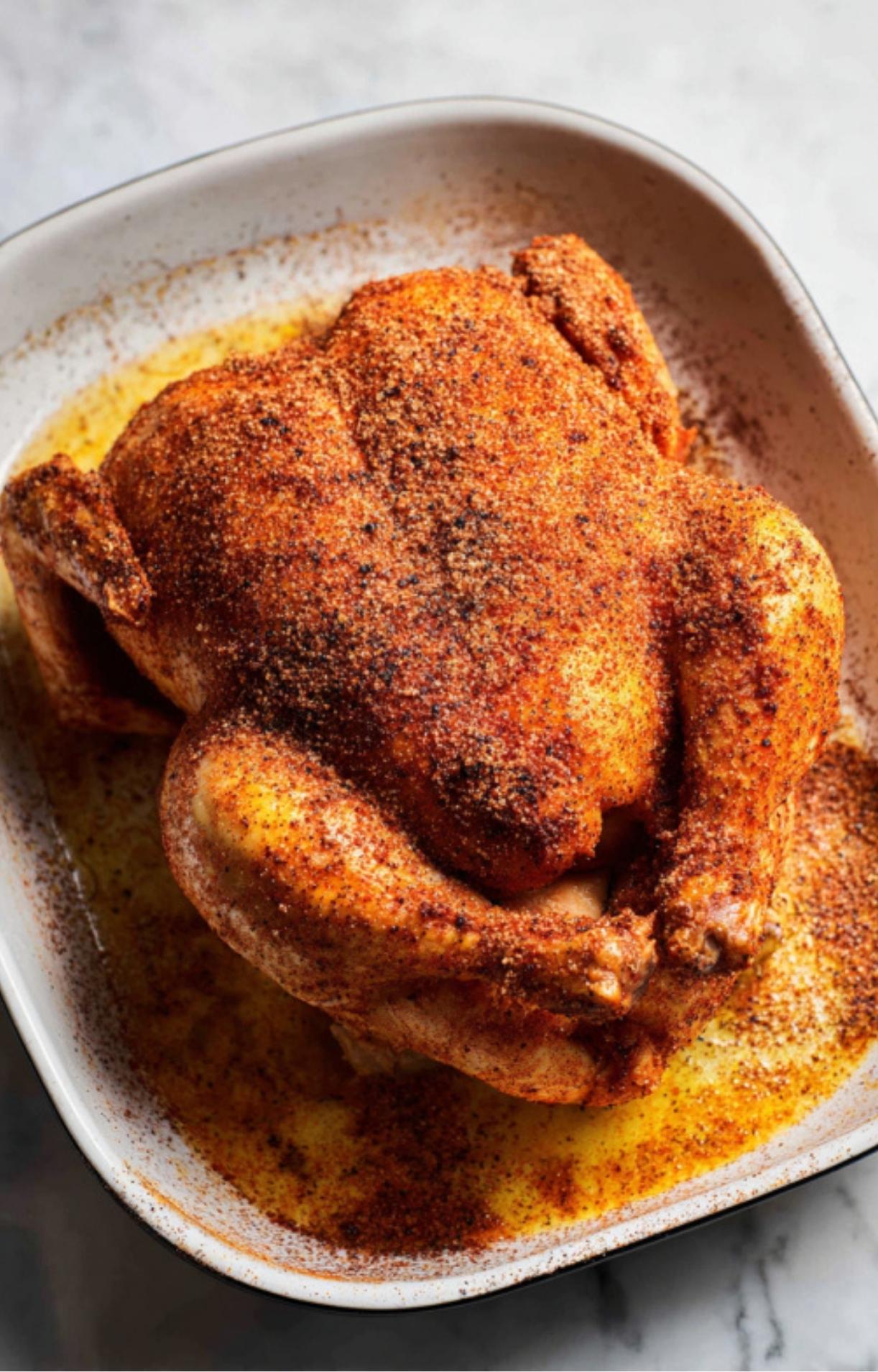 A raw whole chicken resting in a white ceramic baking dish, generously coated in a thick layer of reddish-brown dry rub.