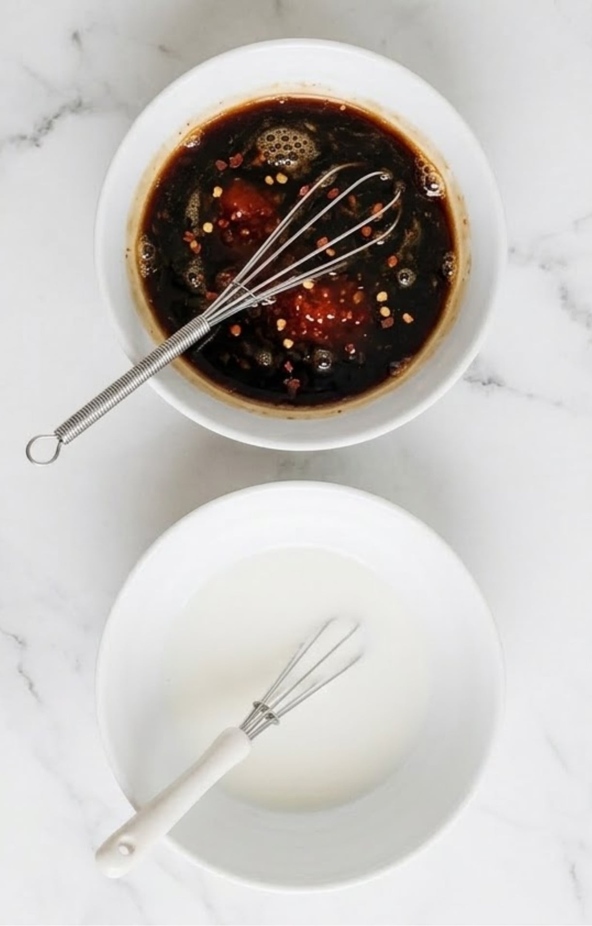 Two white bowls with whisks, one containing a dark savory sauce with red chili flakes and the other a white cornstarch slurry for thickening Beijing beef and broccoli.