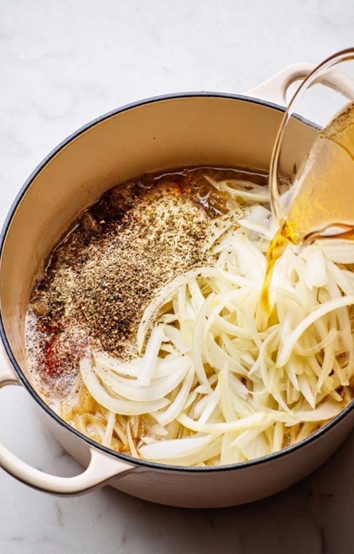Sliced white onions and seasonings in a Dutch oven with beer being poured over them to prepare beer brats on grill.