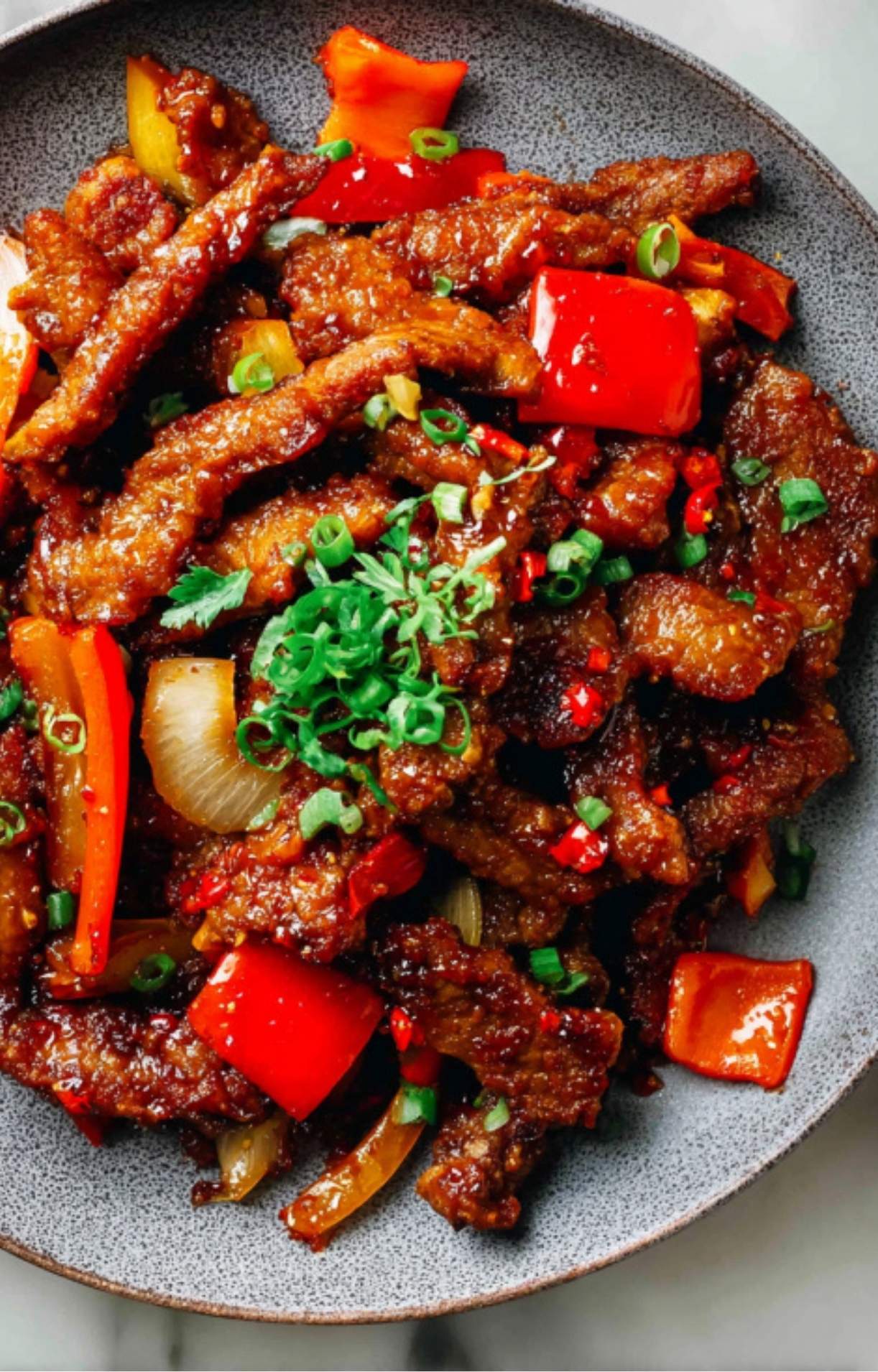 A close-up, top-down view of the crispy beef and peppers on a textured grey plate, garnished with scallions and red chilies.