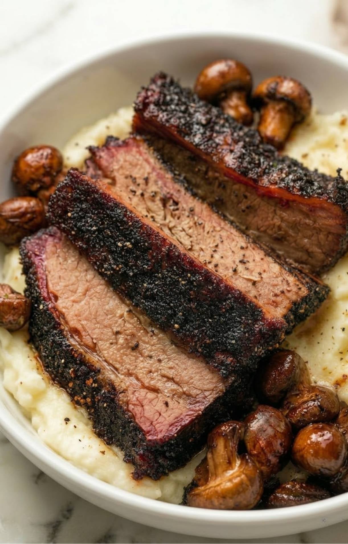 Close-up of brisket slices nestled in creamy mashed potatoes and roasted mushrooms in a white bowl.