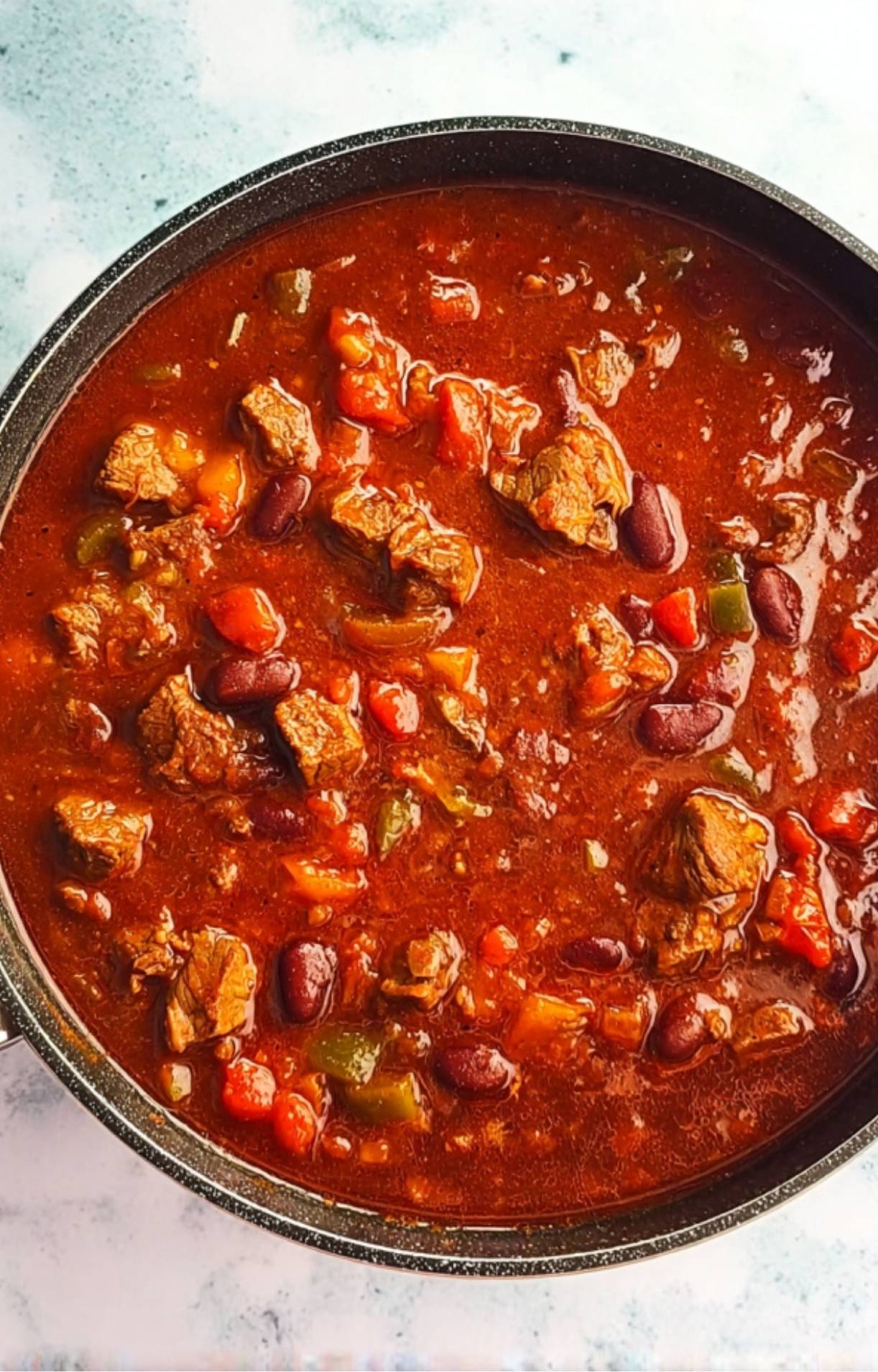 A wide view of Smoked Brisket Chili bubbling in a pot, showing a rich tomato-based broth with visible beans, beef, and peppers.