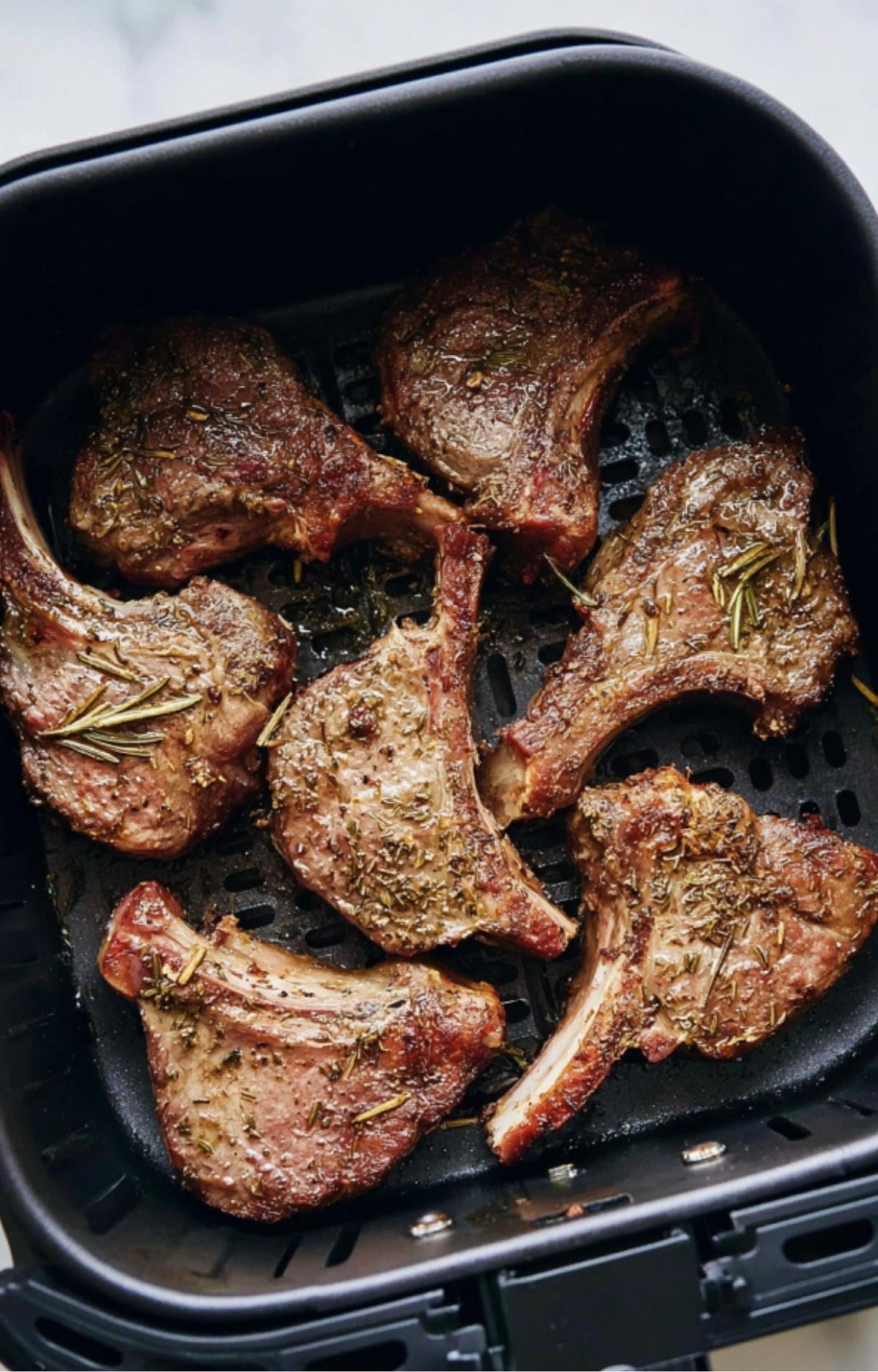 Well-browned lamb chops seasoned with dried rosemary and thyme sitting in an air fryer basket after cooking.