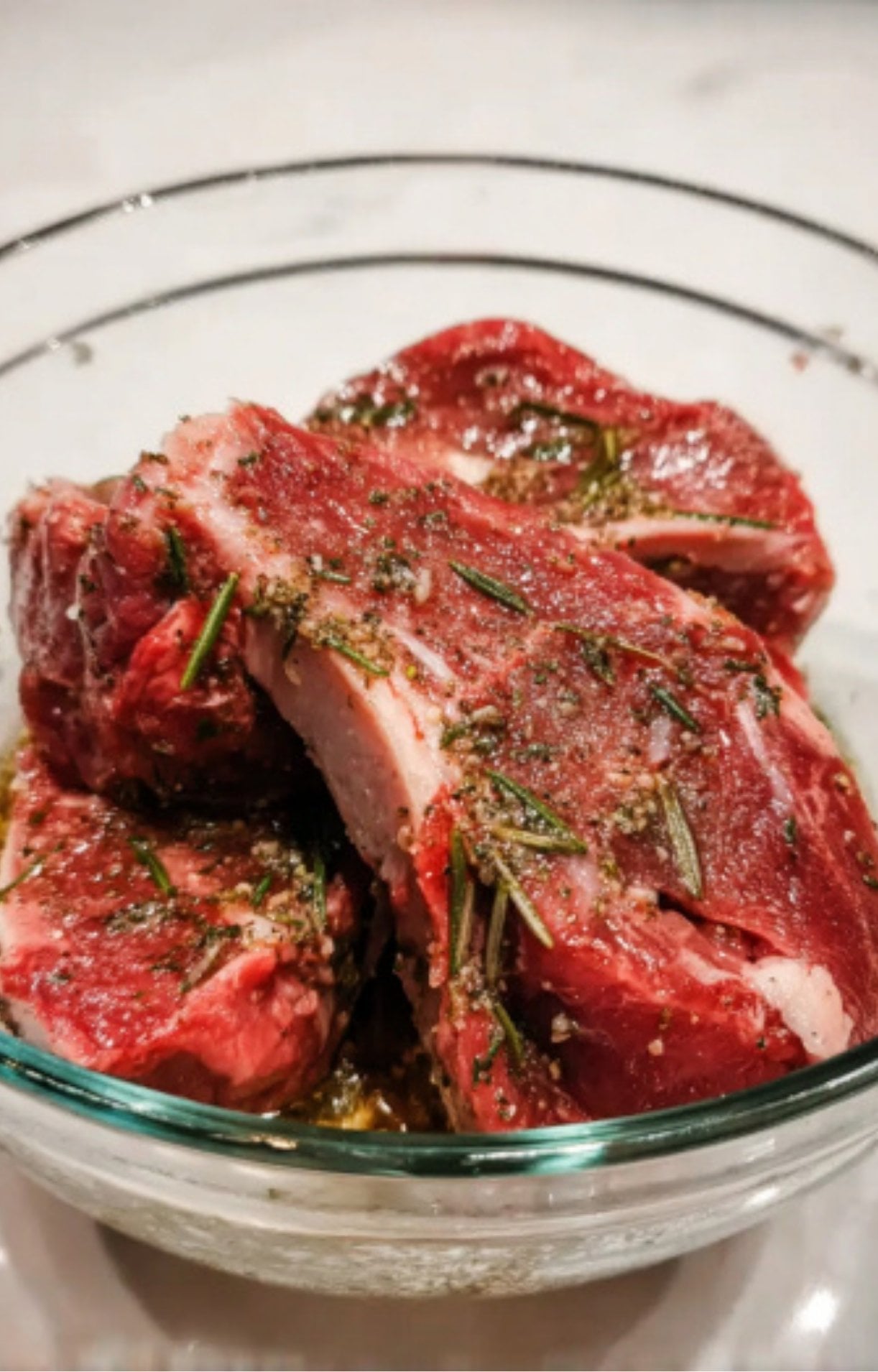 Raw lamb leg steaks being coated in a herb and garlic marinade within a clear glass mixing bowl.