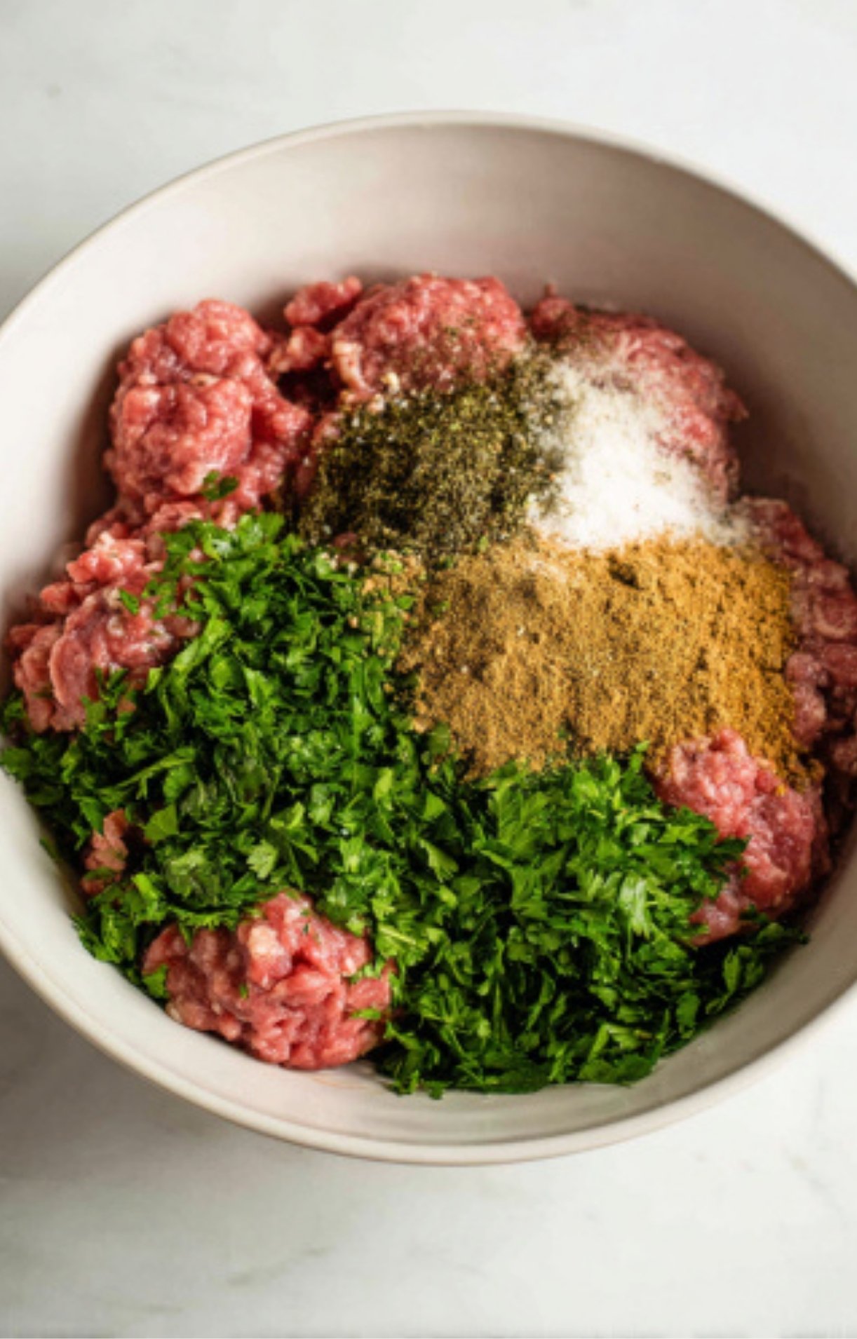 A large mixing bowl containing ground lamb topped with fresh parsley, cumin, coriander, and garlic.