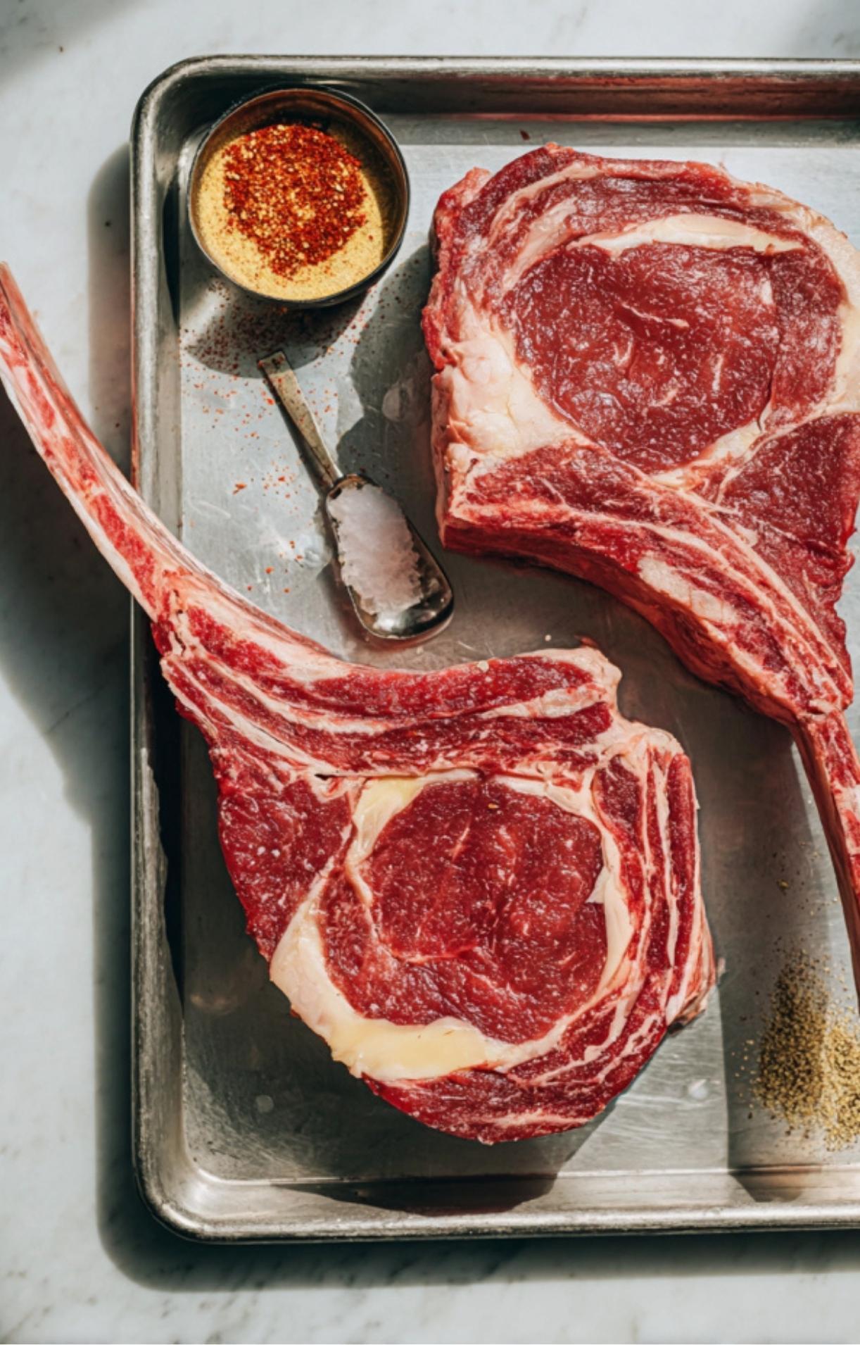 Two large, raw tomahawk ribeye steaks resting on a silver baking sheet next to a small bowl of dry rub seasoning and flaky sea salt.