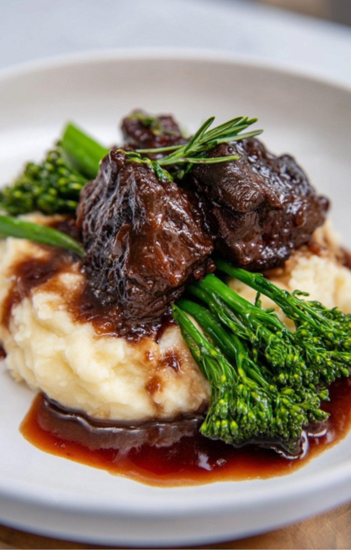 A serving of tender Wagyu Beef Cheeks Recipe over creamy mashed potatoes with a side of broccolini.