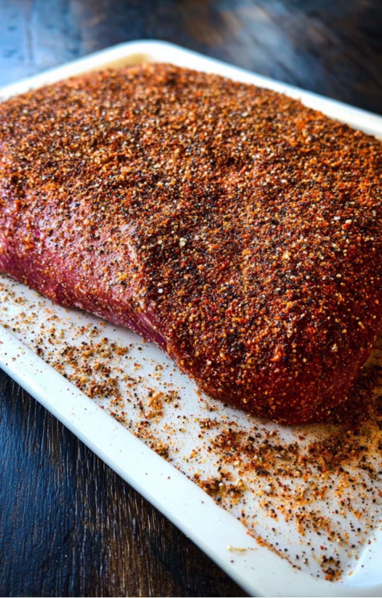 A close-up shot of a raw beef brisket being heavily coated with a savory dry rub on a white prep tray.