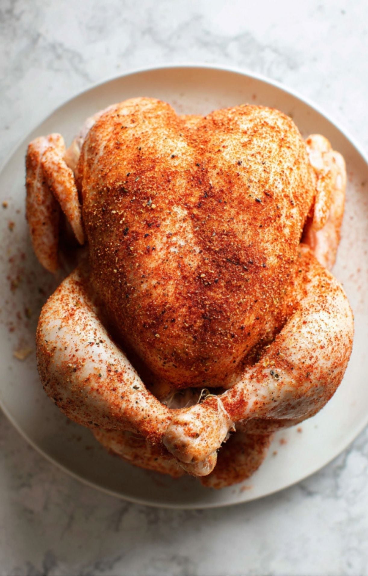 A top-down shot of a raw whole chicken generously coated in a red-orange spice rub, resting on a white plate with visible specks of seasoning on the plate's edge.