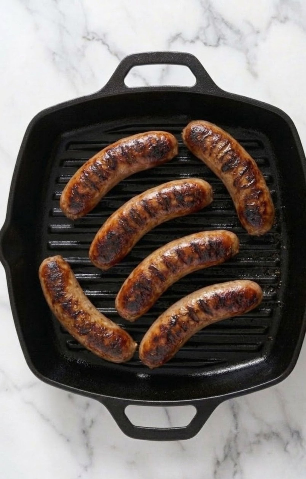 Six juicy bratwurst sausages being seared on a black cast iron grill pan with distinct char marks for a beer brats on grill recipe.