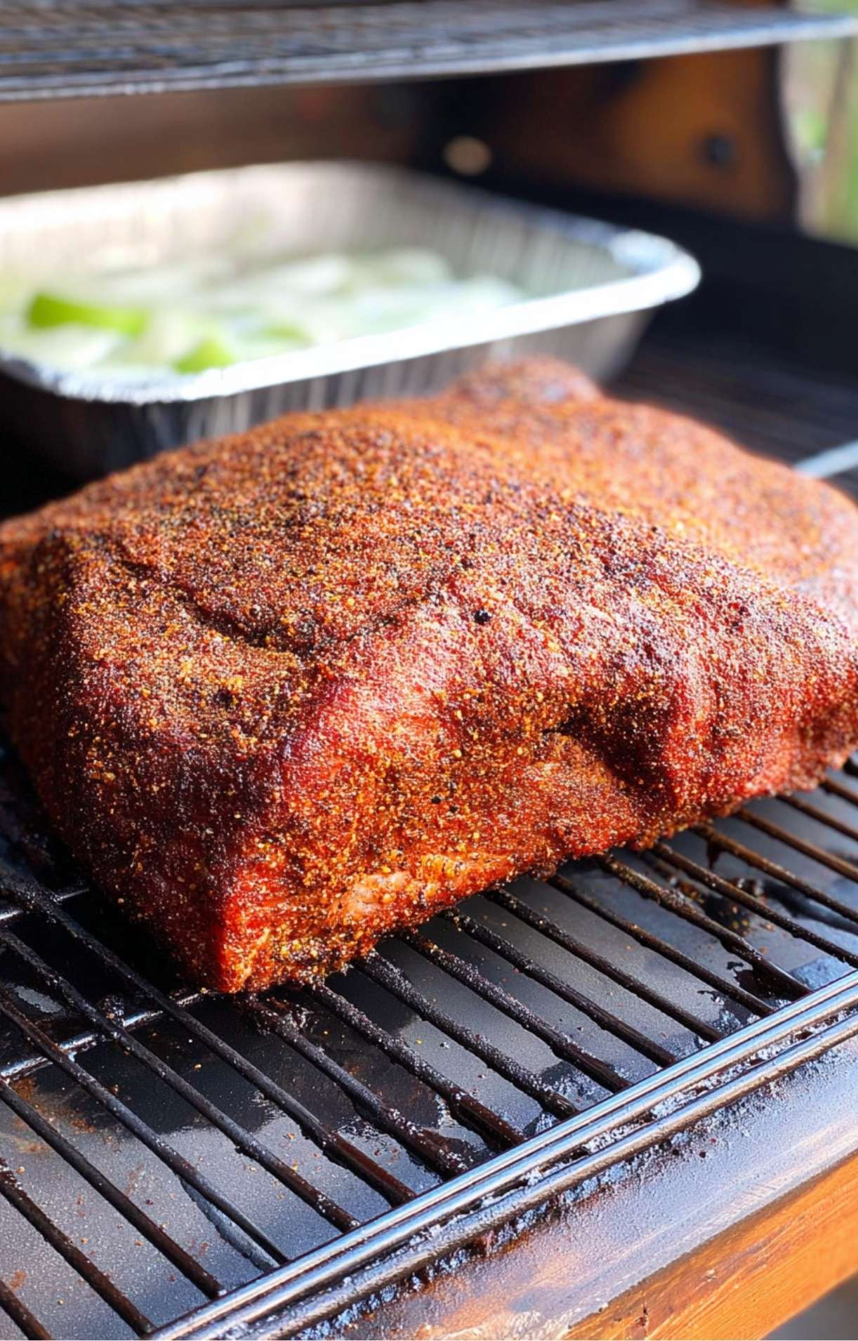 Seasoned brisket cooking on grill grates with a pan of water in the background for moisture.