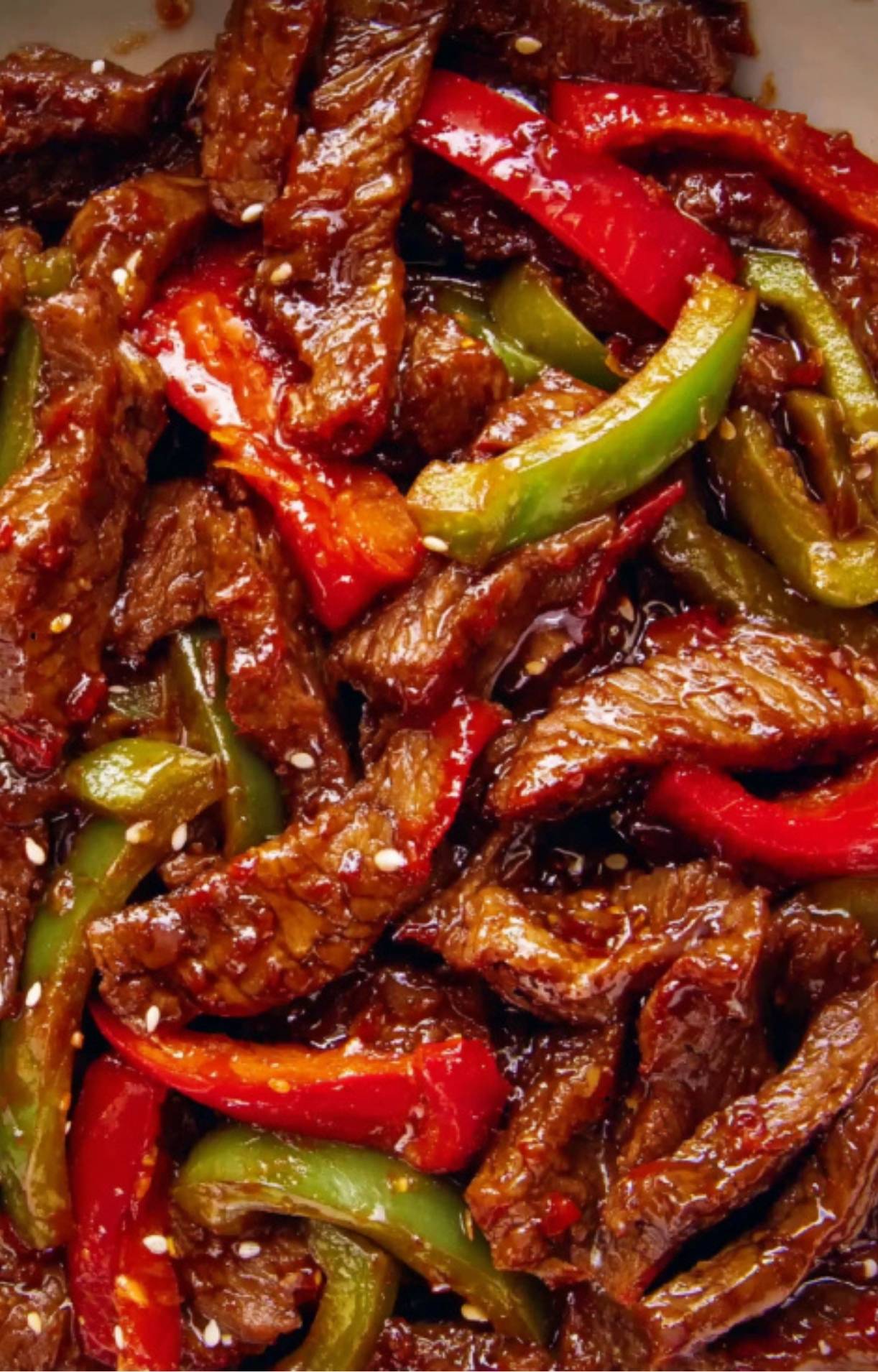 A macro shot of cooked Beijing Beef and bell peppers thoroughly coated in a dark, shimmering Beijing Beef sauce and sprinkled with sesame seeds.