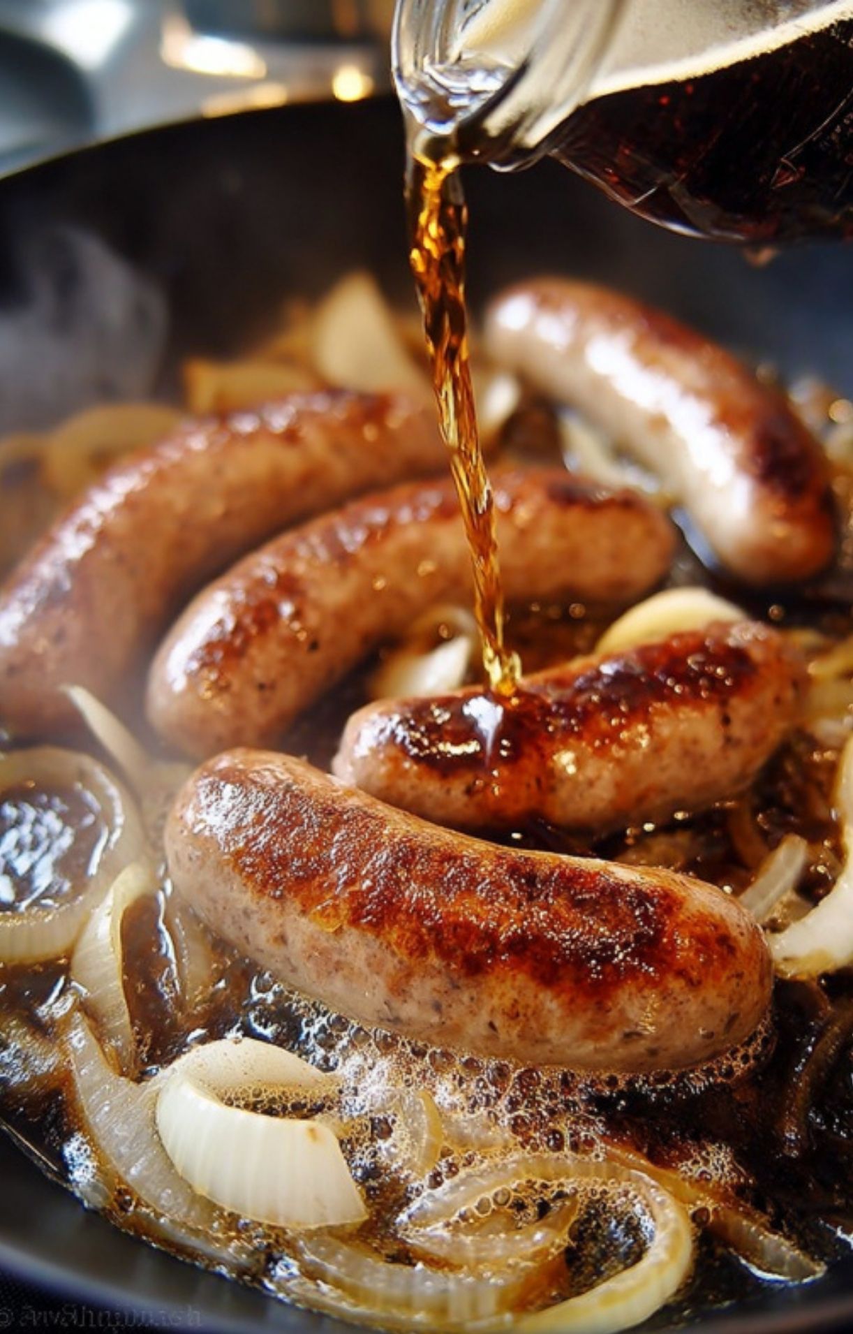 Dark beer being poured from a glass bottle over seared bratwurst sausages and sliced onions in a skillet.
