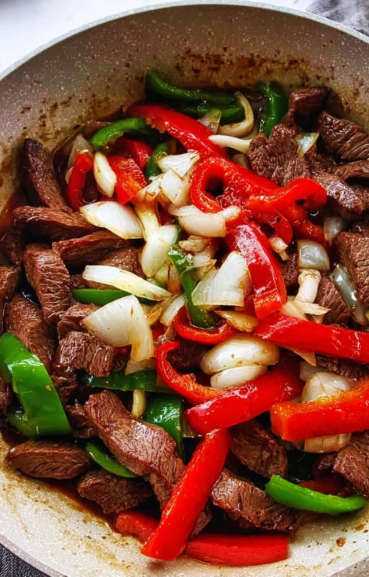 Thinly sliced beef, red and green bell peppers, and white onions being stir-fried together in a large light-colored skillet.