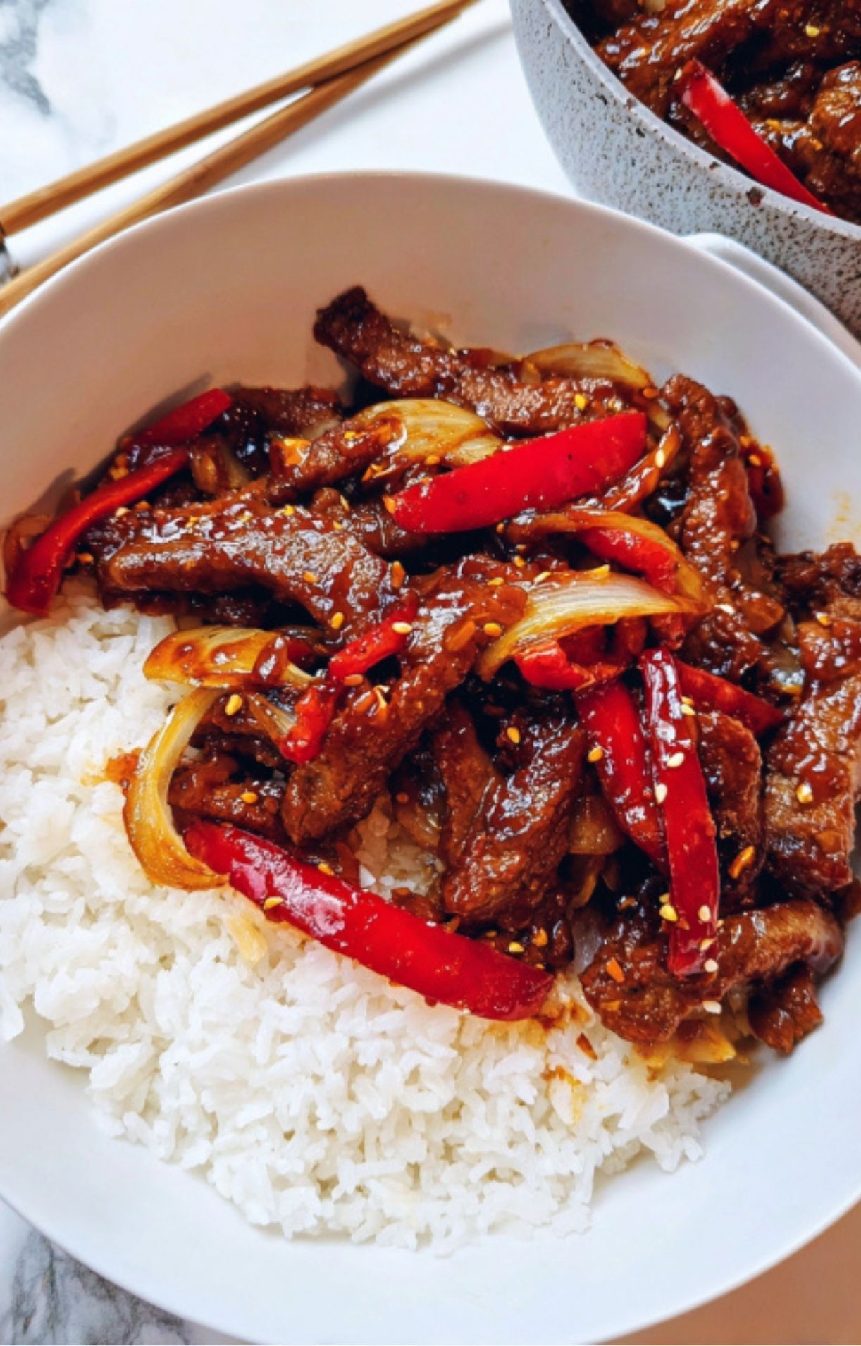 A white bowl filled with a generous serving of fluffy white rice topped with glossy Beijing Beef strips, red bell peppers, and onions, garnished with sesame seeds.