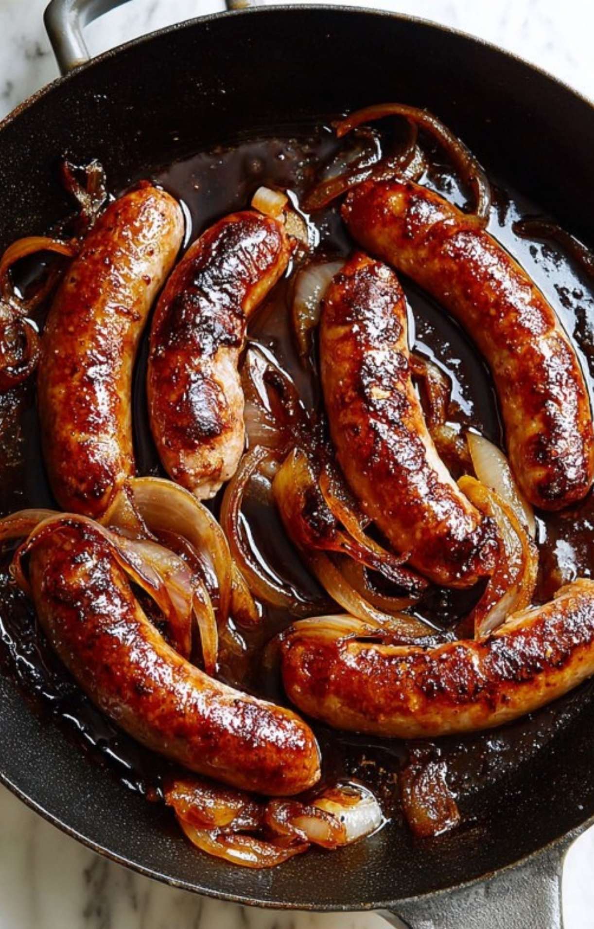 Finished beer brats with deeply caramelized onions in a dark, reduced beer glaze in a cast iron pan.