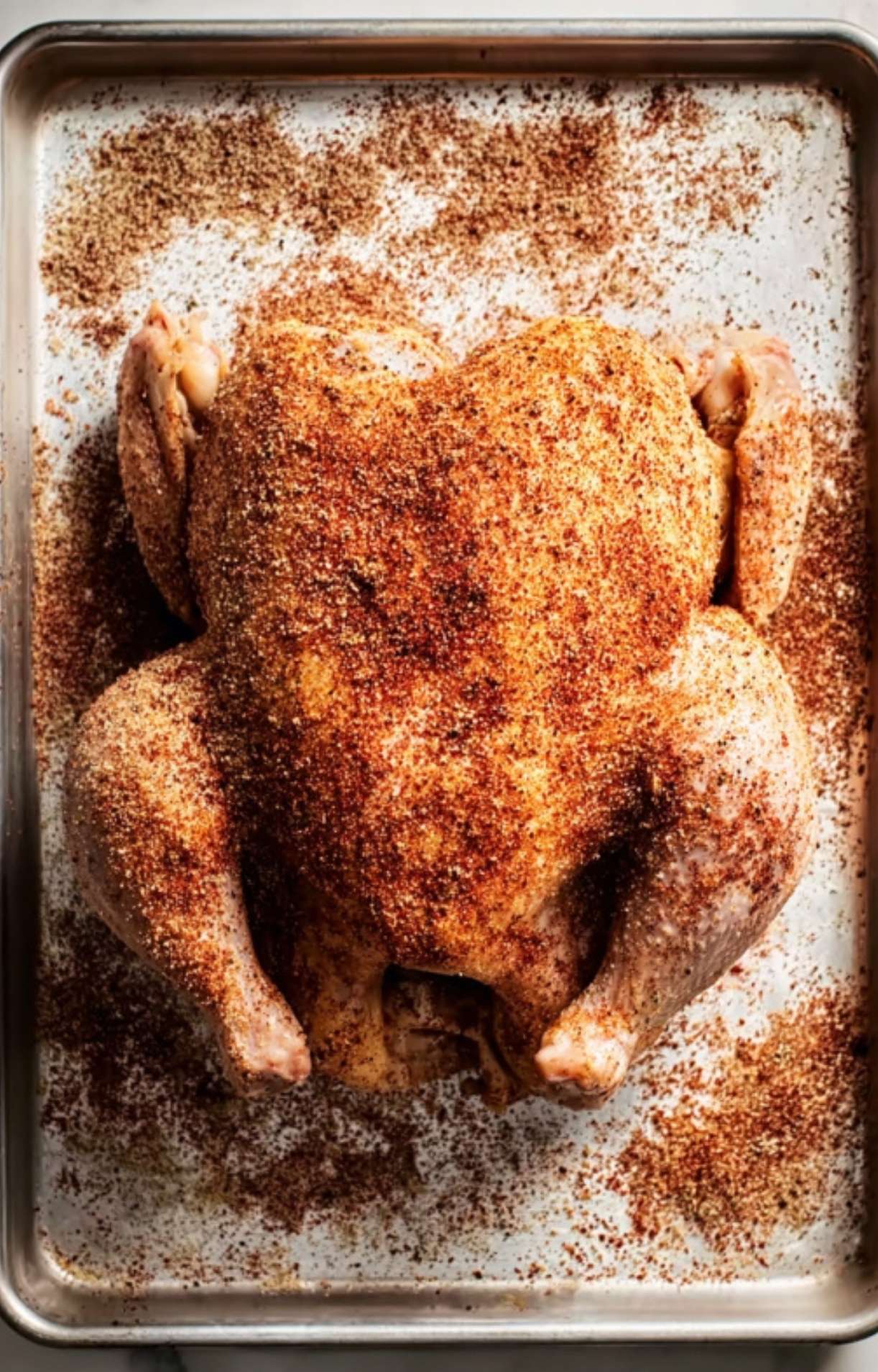 A whole raw chicken placed on a baking sheet, generously coated from top to bottom with a thick layer of reddish-brown dry rub.