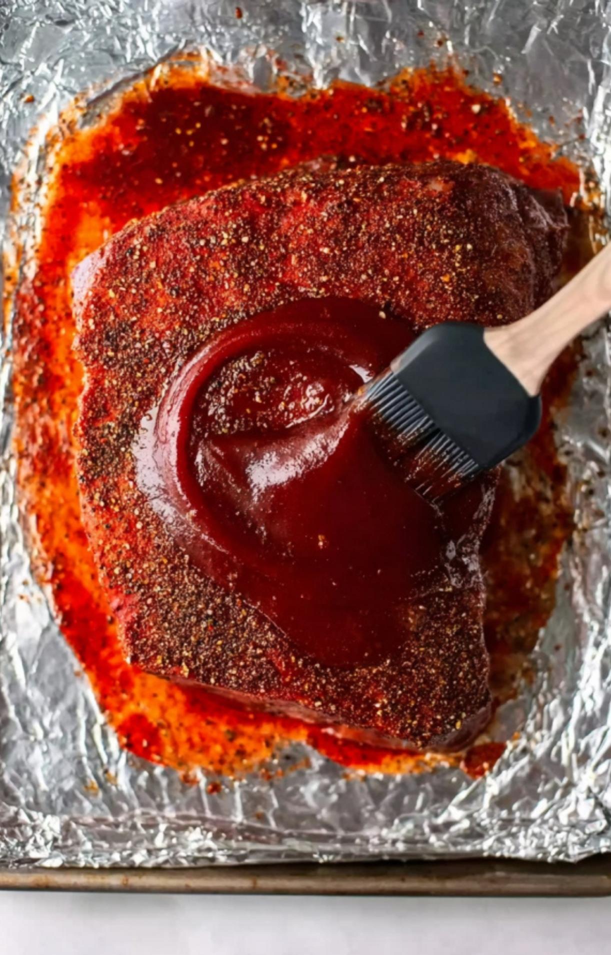 Using a silicone brush to spread a thick, dark red barbecue sauce over the top of the seasoned beef brisket.