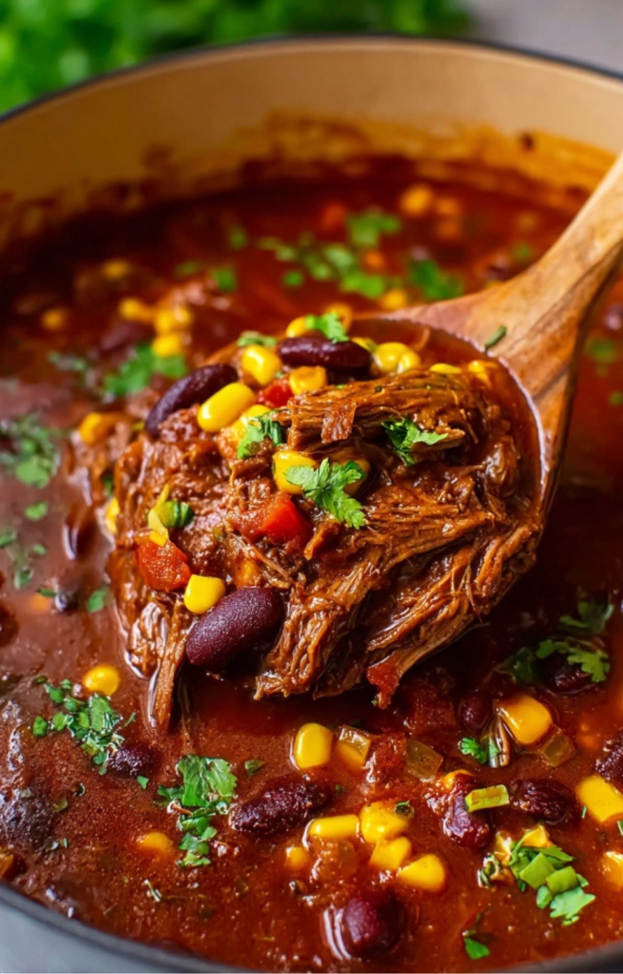 A wooden spoon lifting a generous serving of Smoked Brisket Chili, highlighting the shredded texture of the smoked beef mixed with corn and beans.