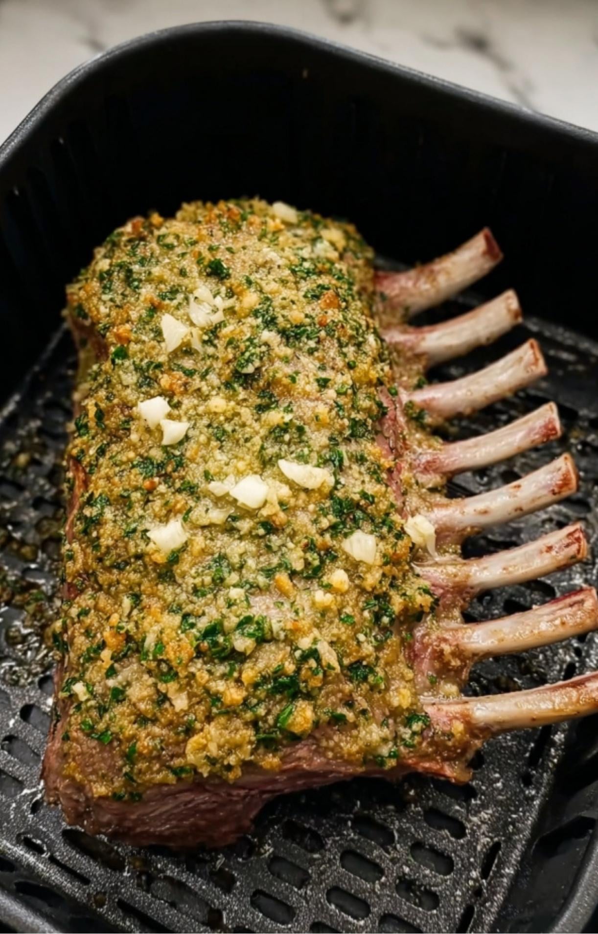 A rack of lamb in the air fryer basket topped with a thick layer of green herbs and minced garlic pieces.