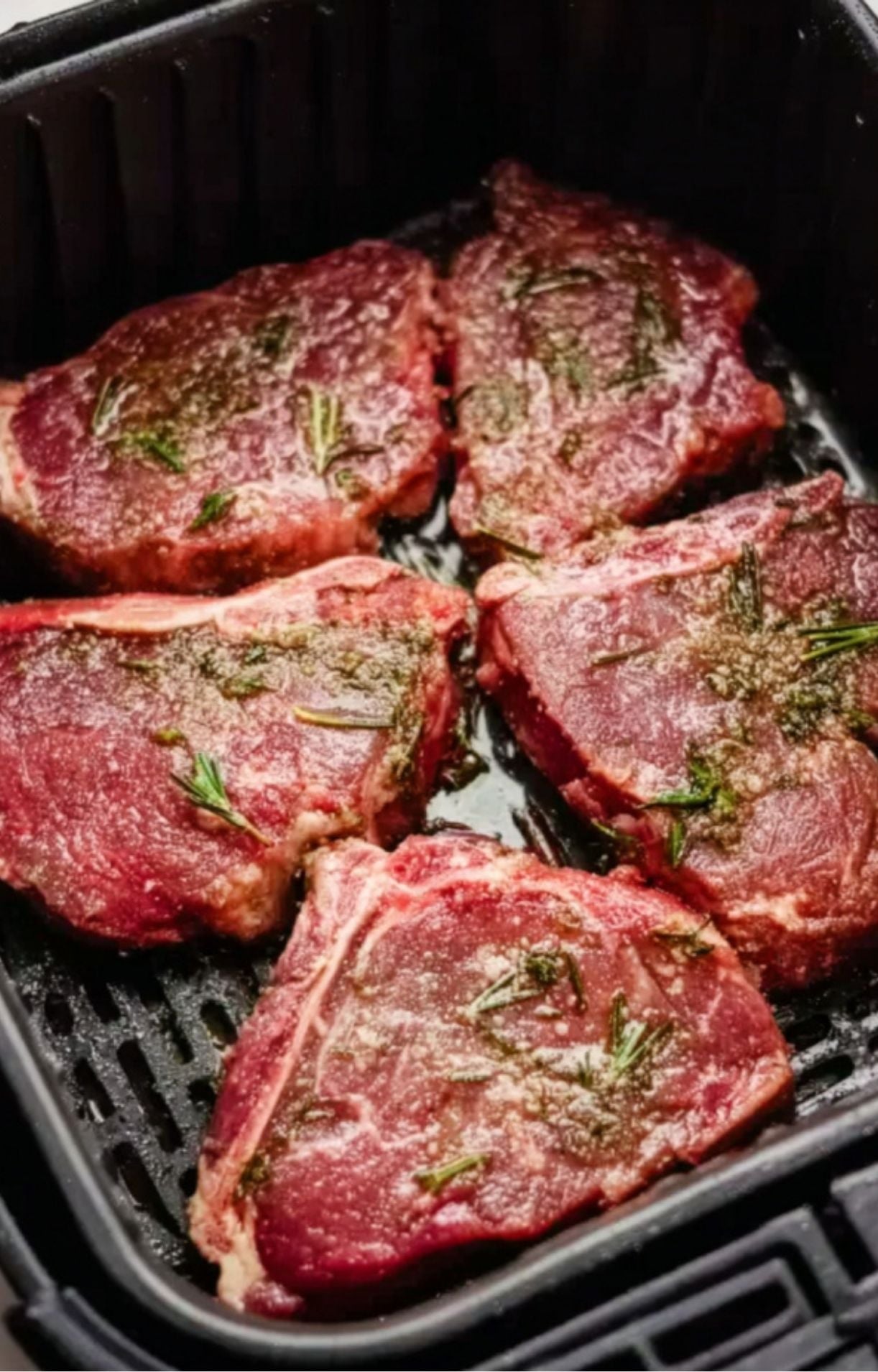 Four seasoned raw lamb steaks placed in a single layer inside a black air fryer basket ready for cooking.