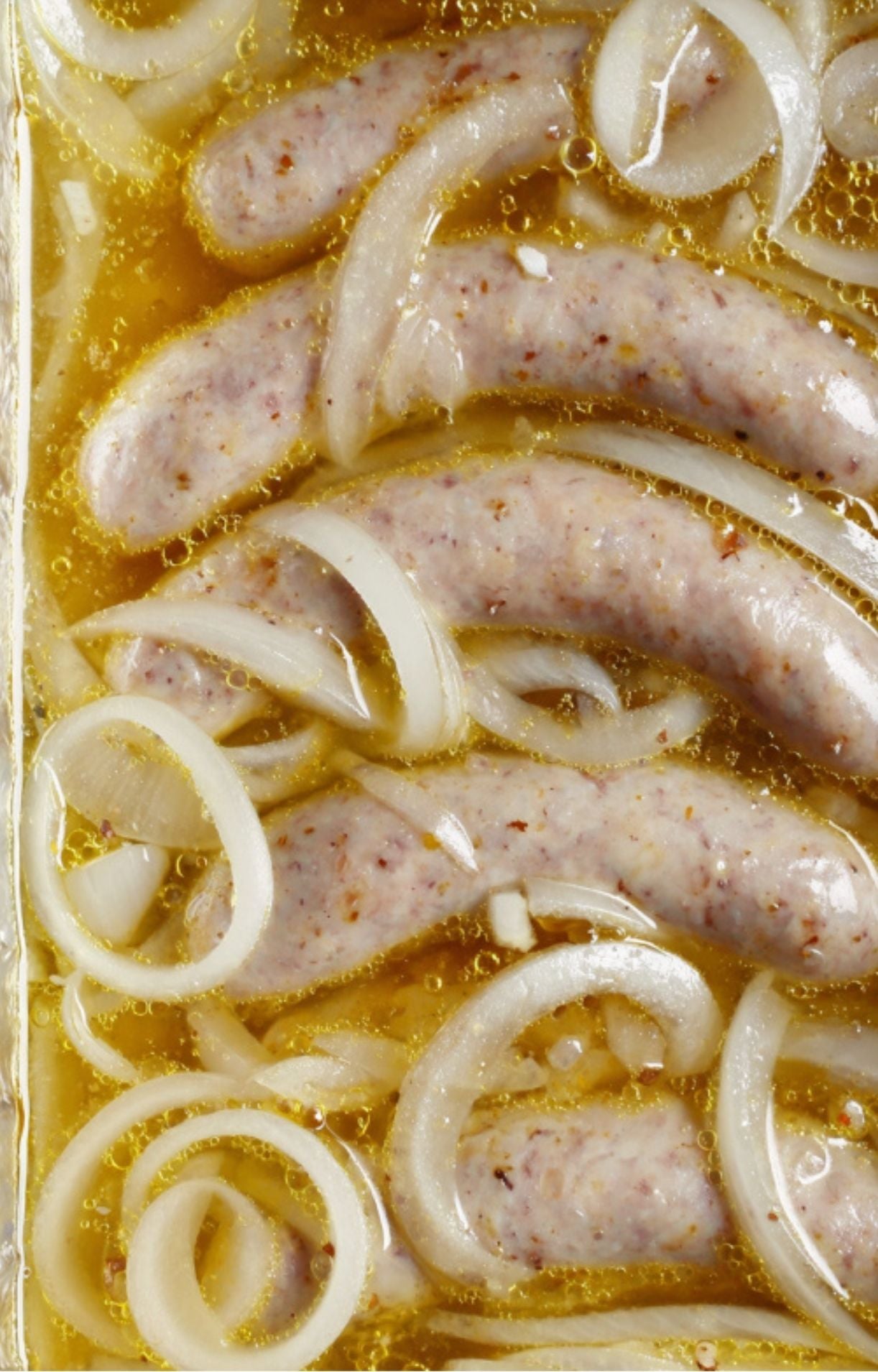 Close-up of bratwurst sausages simmering in a golden beer and onion broth within a glass baking dish.