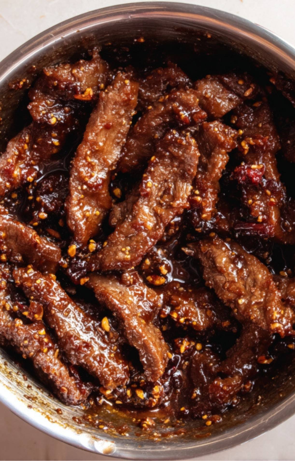 Crispy cooked beef strips being thoroughly coated in a thick, glossy dark sauce in a metal mixing bowl.