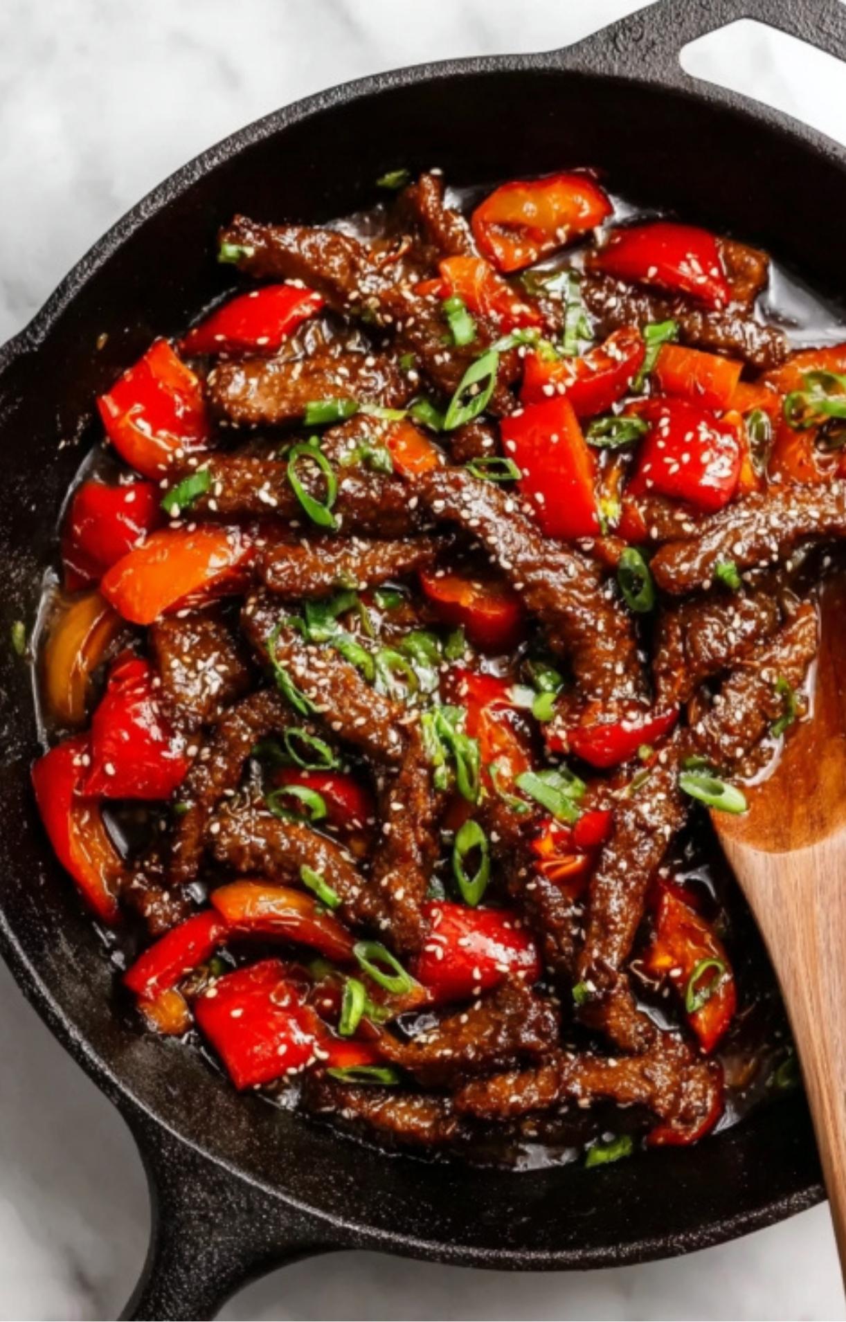 Crispy beef and vibrant red bell peppers simmering in a dark savory sauce in a cast-iron skillet, topped with green onions.