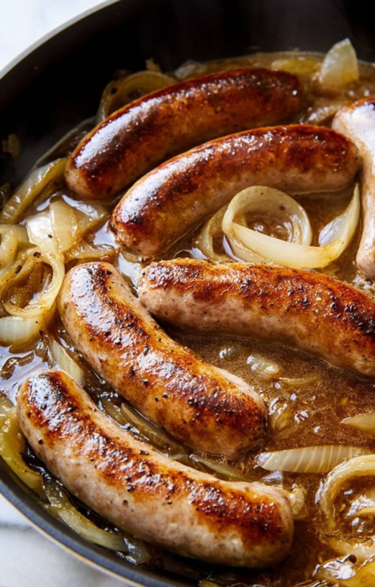 Bratwurst sausages simmering in a beer and onion liquid base in a large skillet.