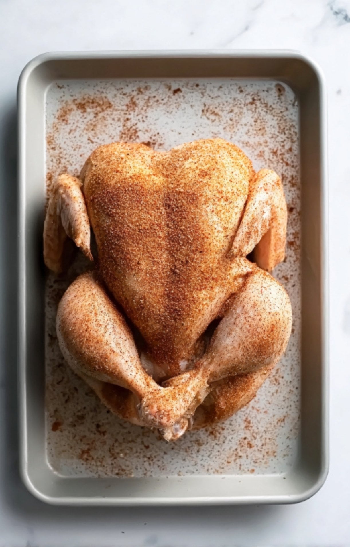 A whole raw chicken in a pan, generously coated with a reddish-brown spice rub over its entire surface.