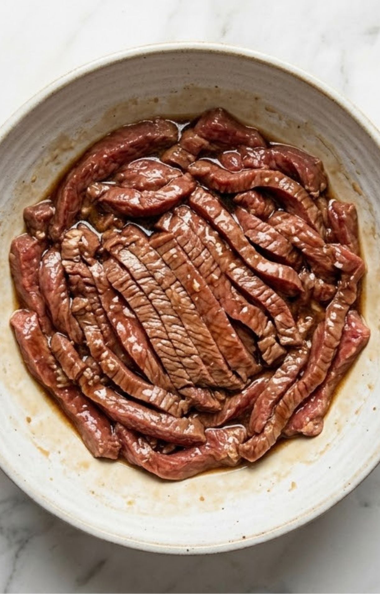 Thinly sliced strips of raw beef laid out in a white bowl, lightly coated in a liquid marinade to tenderize and flavor the meat before cooking.