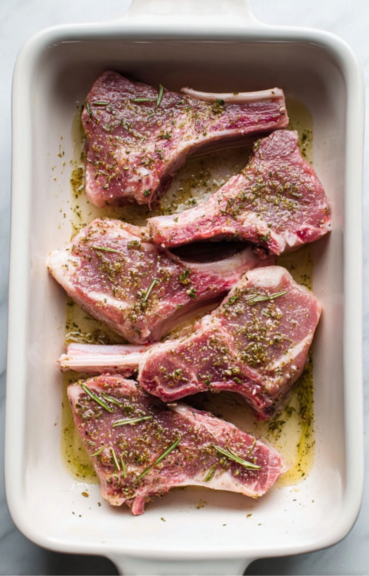 Five raw lamb chops marinating in a white ceramic dish with olive oil, salt, pepper, and fresh rosemary sprigs.