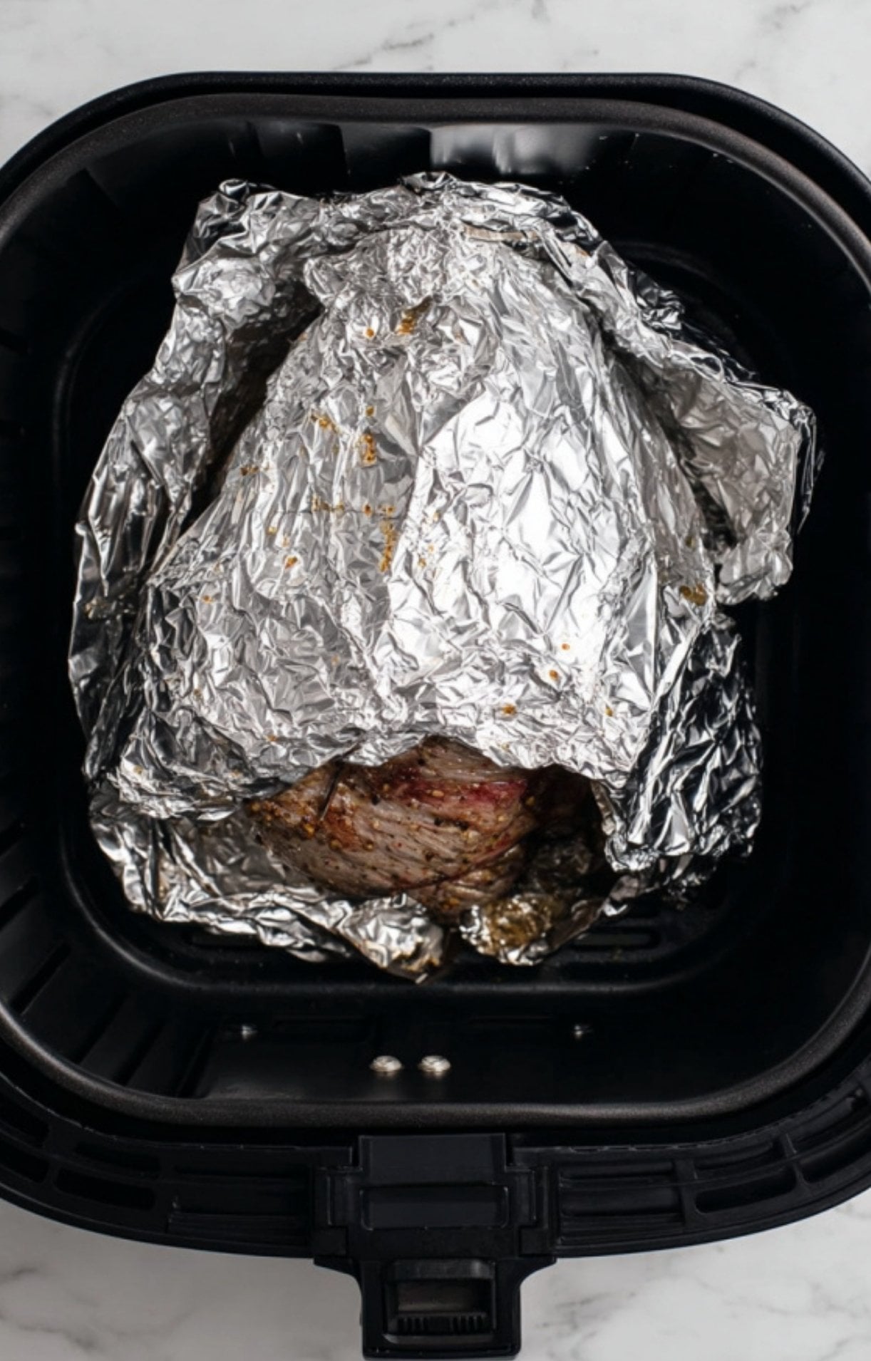 A marinated leg of lamb partially wrapped in aluminum foil inside a black air fryer basket to ensure even cooking.