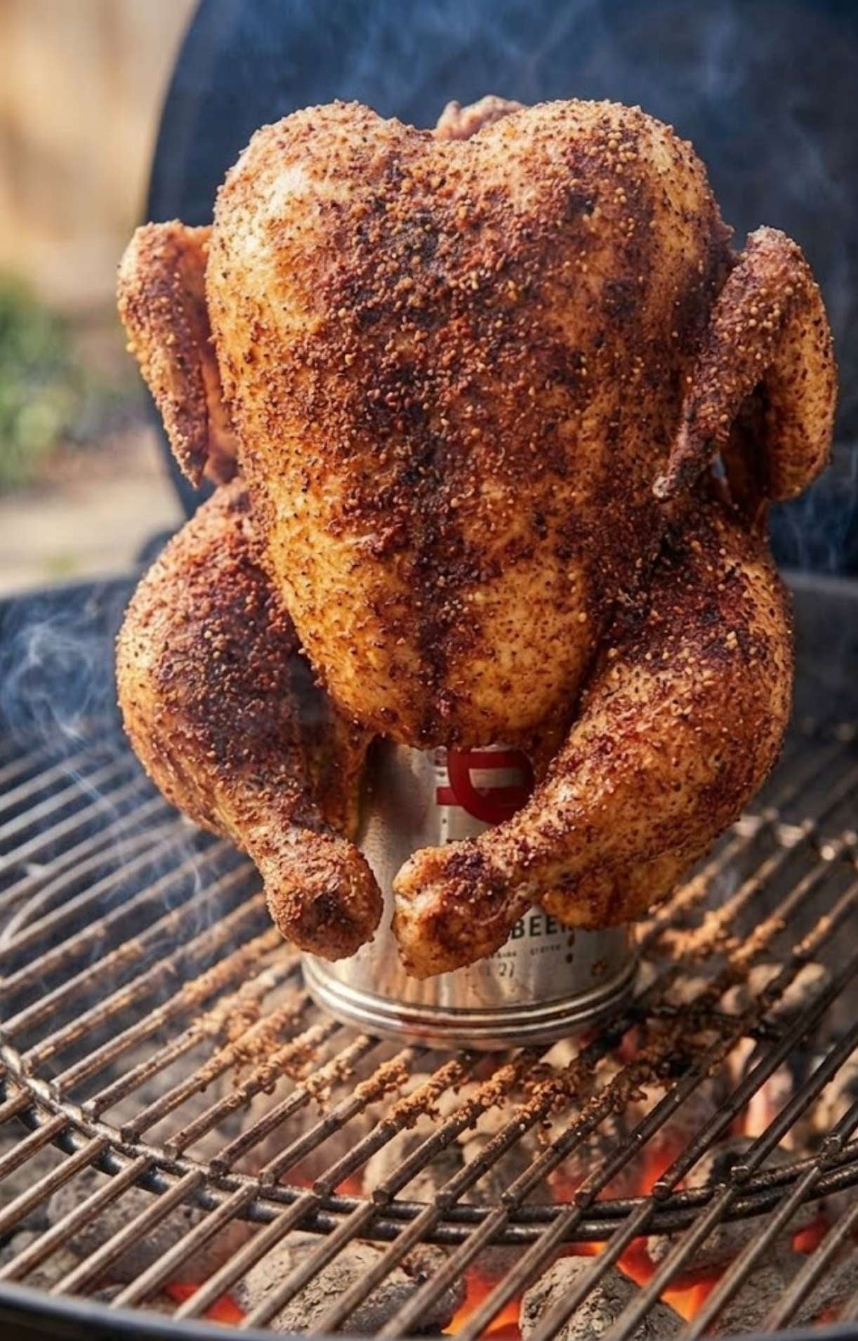 A dry-rubbed chicken cooking vertically on a beer can over hot charcoal briquettes in a kettle grill for Weber Beer Can Chicken.